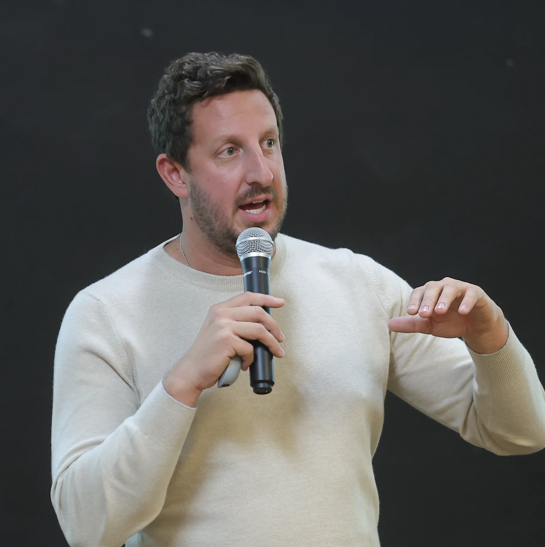 Man in beige sweater speaking into a microphone with hand gesturing against a black background.