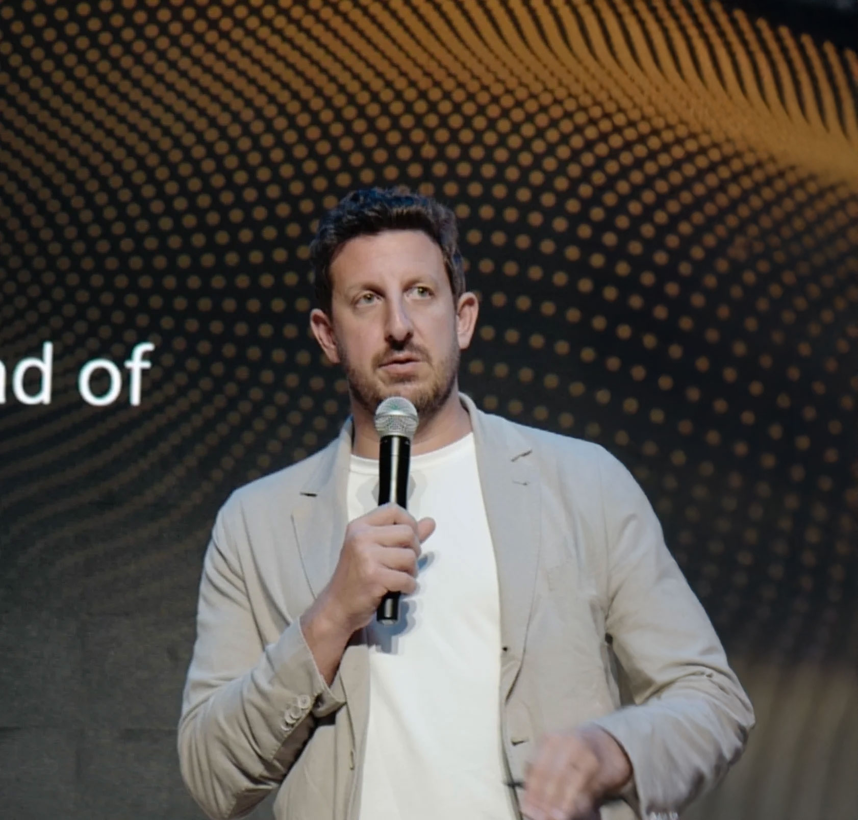 Man with short dark hair and beard speaking into a microphone, wearing a beige blazer and white shirt against a dark patterned background.