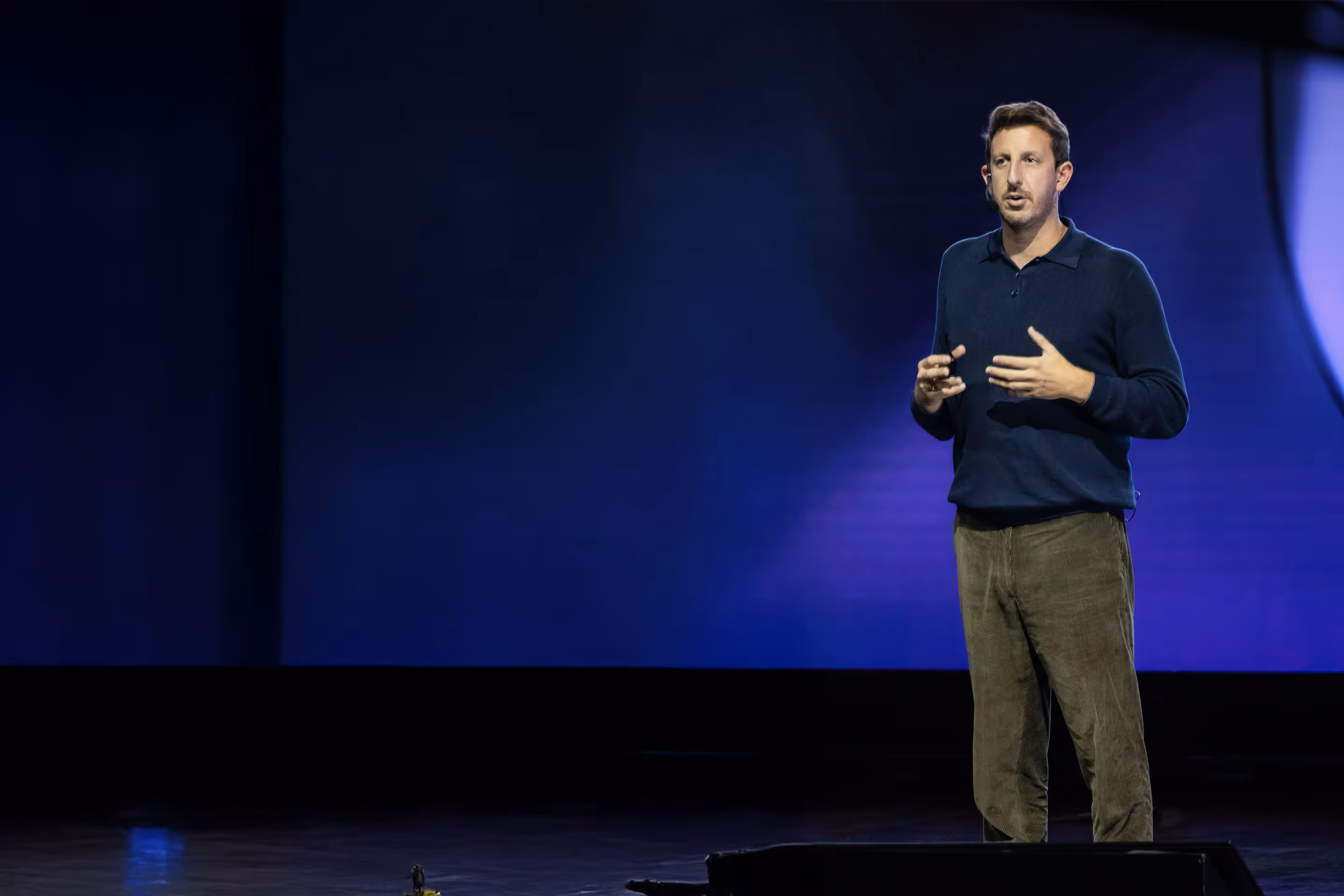 Man in dark sweater speaking and gesturing during a presentation with large letters 'SE' in the background.
