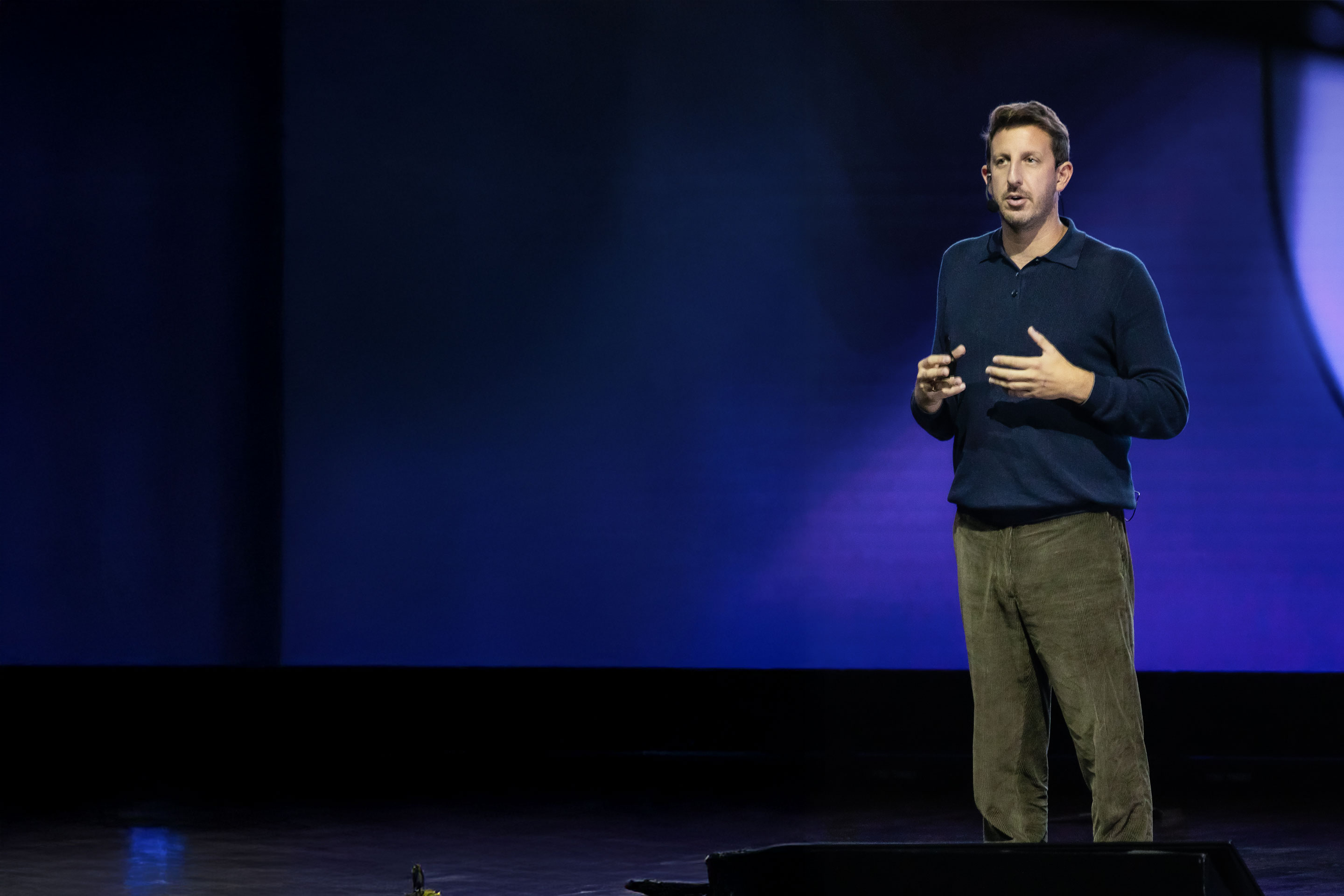 Man in dark sweater speaking and gesturing during a presentation with large letters 'SE' in the background.