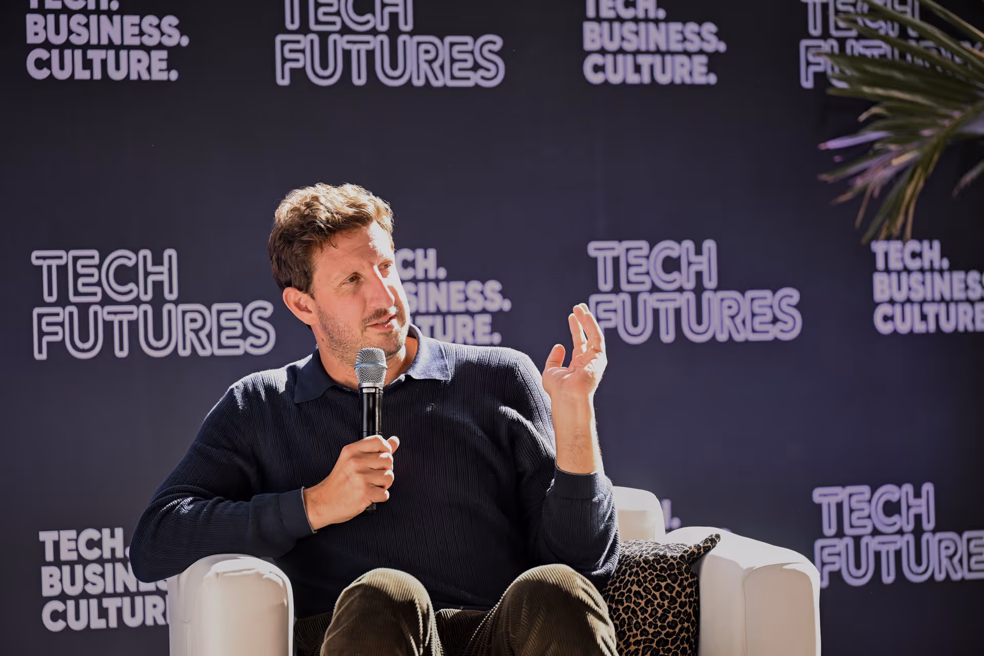 A man in a dark sweater holding a microphone and gesturing while seated on a white chair with a leopard print pillow against a backdrop with the text 'Tech Futures' and 'Tech. Business. Culture.'
