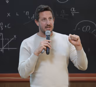Zack Kass in a light sweater speaking into a microphone in front of a blackboard with formulas.