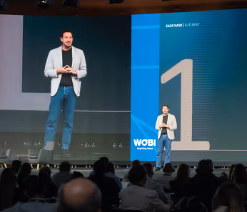 Zack Kass stands on a stage in front of a large screen, speaking into a microphone with his hands clasped together.