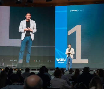 Zack Kass stands on a stage in front of a large screen, speaking into a microphone with his hands clasped together.