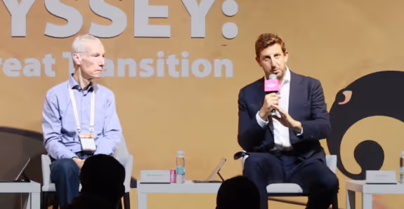 Zack Kass and another man seated on stage during a panel discussion, one speaking into a microphone, with a backdrop featuring partially visible text.