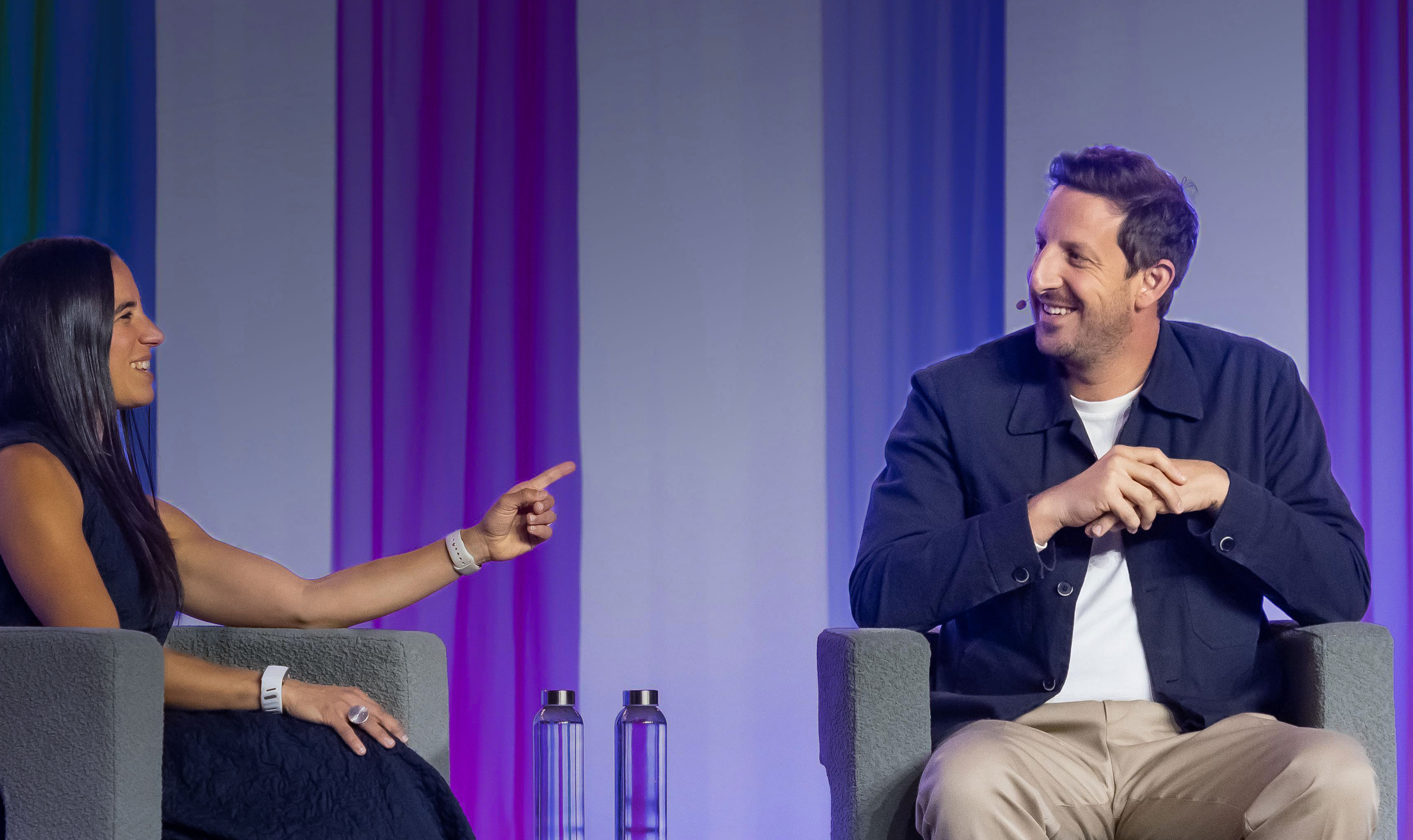 Smiling woman pointing while seated in a gray armchair talking to a smiling Zack Kass wearing a headset microphone seated in another gray armchair.