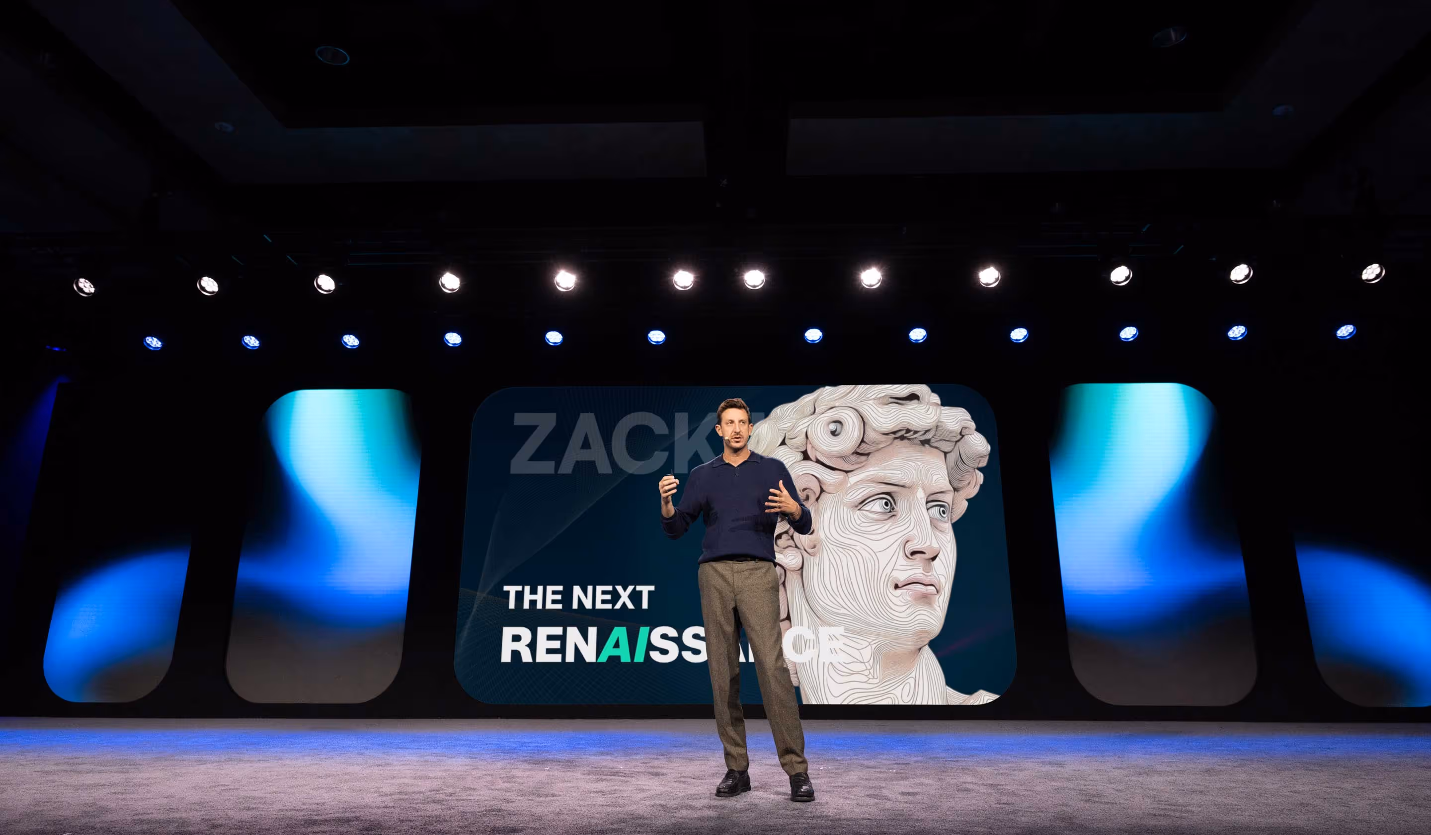 Man giving a presentation on stage with a large screen behind him showing a stylized sculpture head and the words 'The Next Renaissance'.