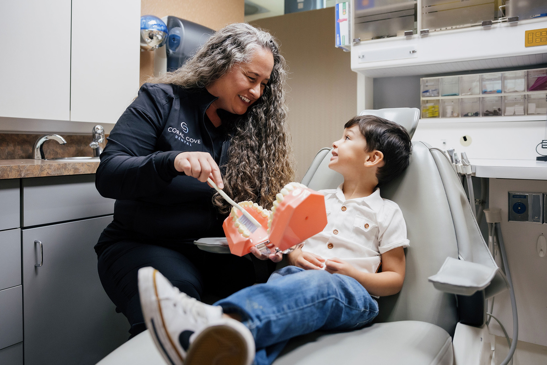Dental professional demonstrating proper brushing technique on a large tooth model to a young boy seated in a dental chair.