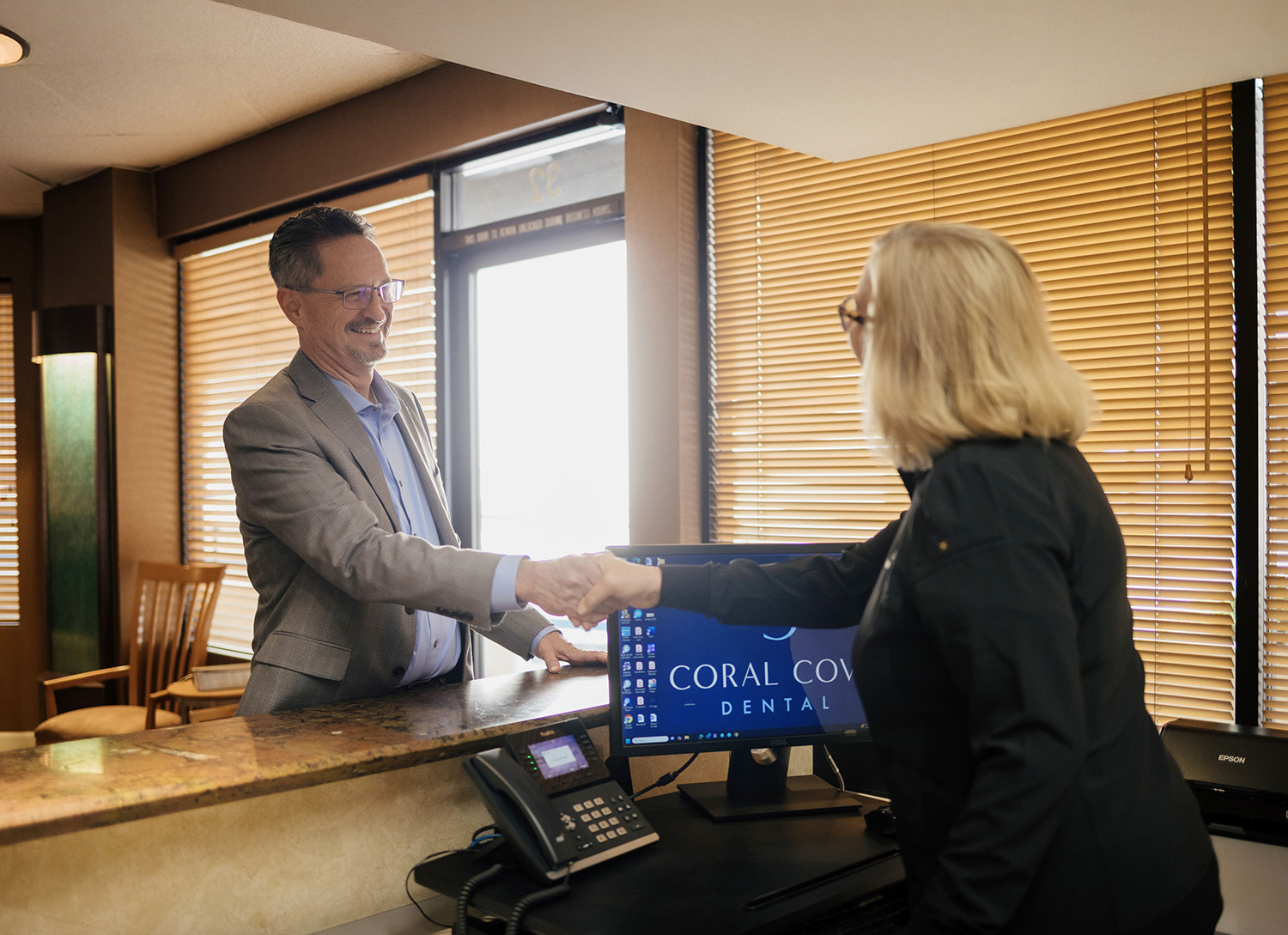 Man and woman shaking hands over a reception desk with a computer screen displaying Coral Cove Dental logo.