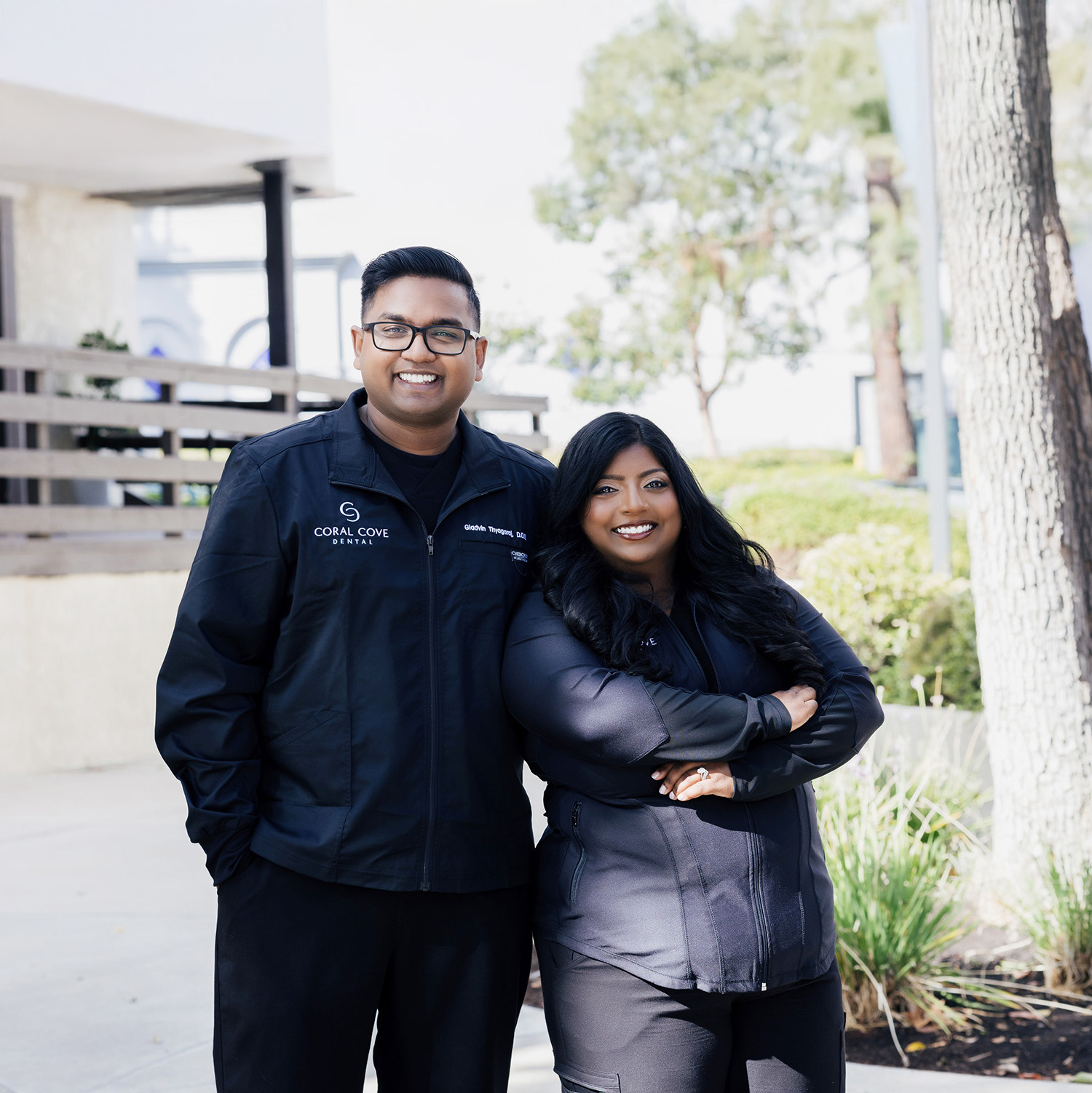 Smiling man and woman wearing Coral Cove Dental jackets standing outdoors in a sunny setting.