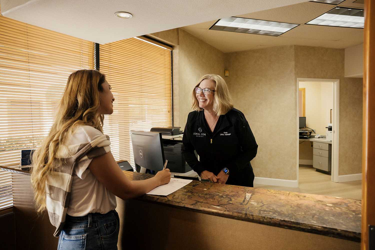 Woman signing in at reception desk while office manager in black jacket smiles behind the counter in a warmly lit office.