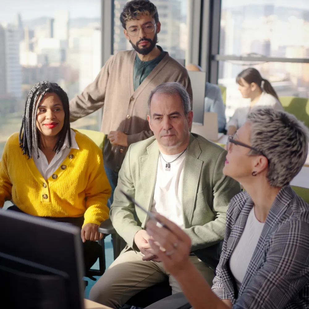 Four diverse colleagues engaged in discussion around a computer in a bright modern office.
