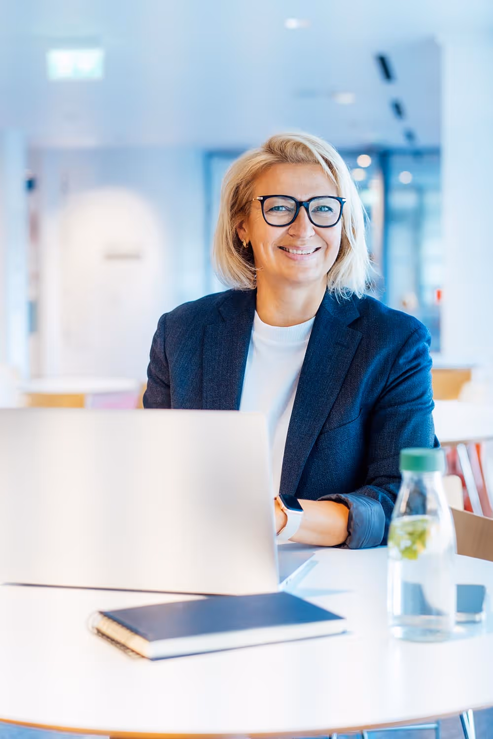 Smiling businesswoman wearing glasses and a navy blazer sitting at a white table with a laptop, notebook, and water bottle.