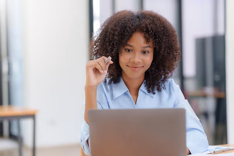 Smiling woman with curly hair in blue shirt working on a laptop in a modern office.
