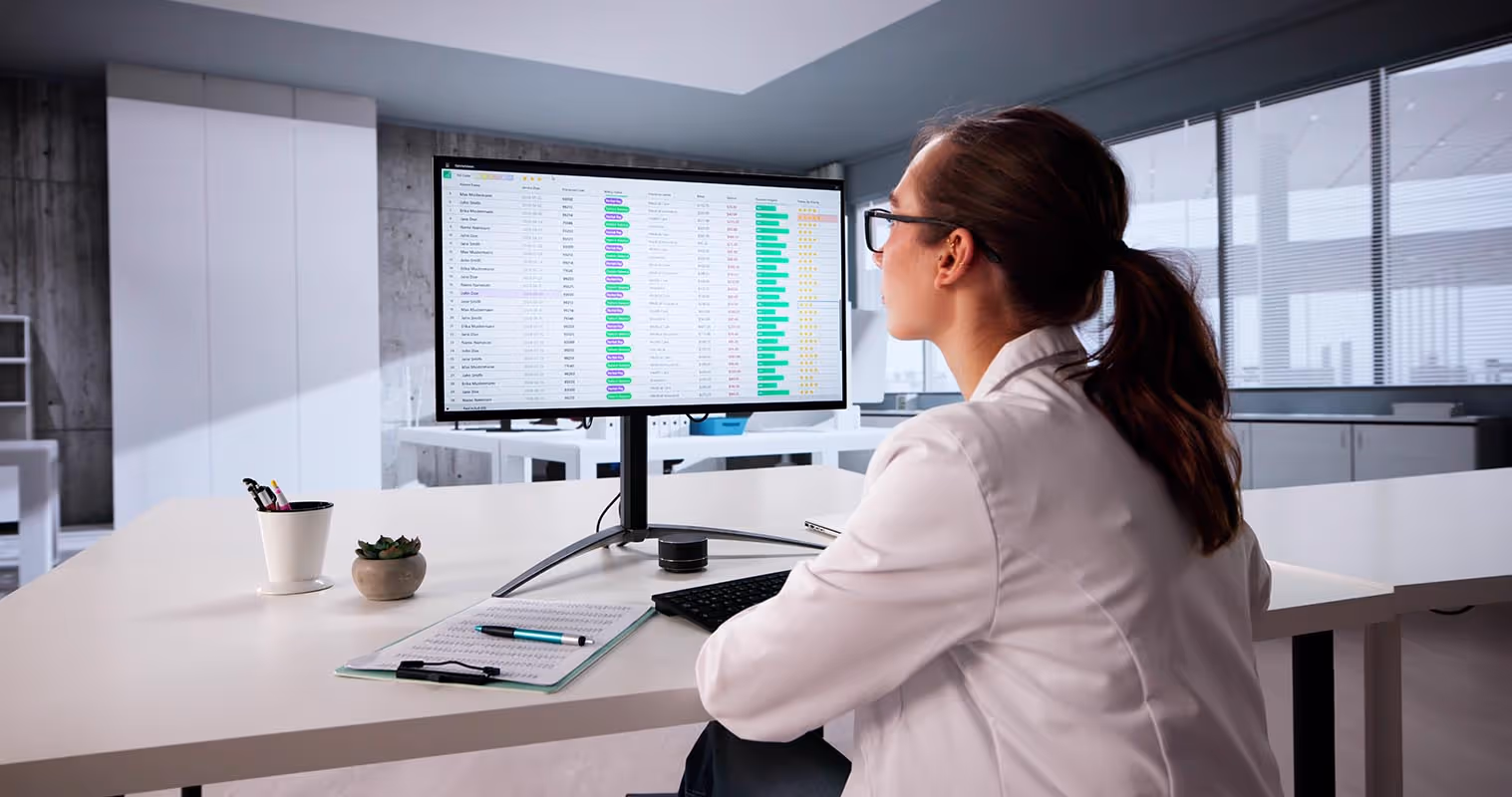 Woman in a white lab coat and glasses analyzing data on a computer monitor in a modern office.