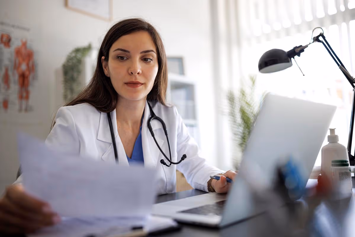 Healthcare worker handling paper documentation while using a laptop, reflecting how Jopari Attach enables accurate, real-time electronic attachment delivery