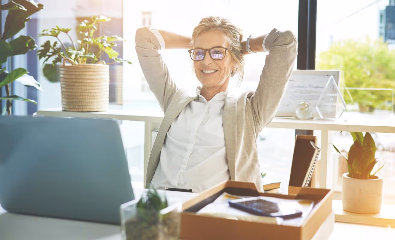Smiling woman in glasses leaning back with hands behind head at a sunlit desk with plants and office supplies.