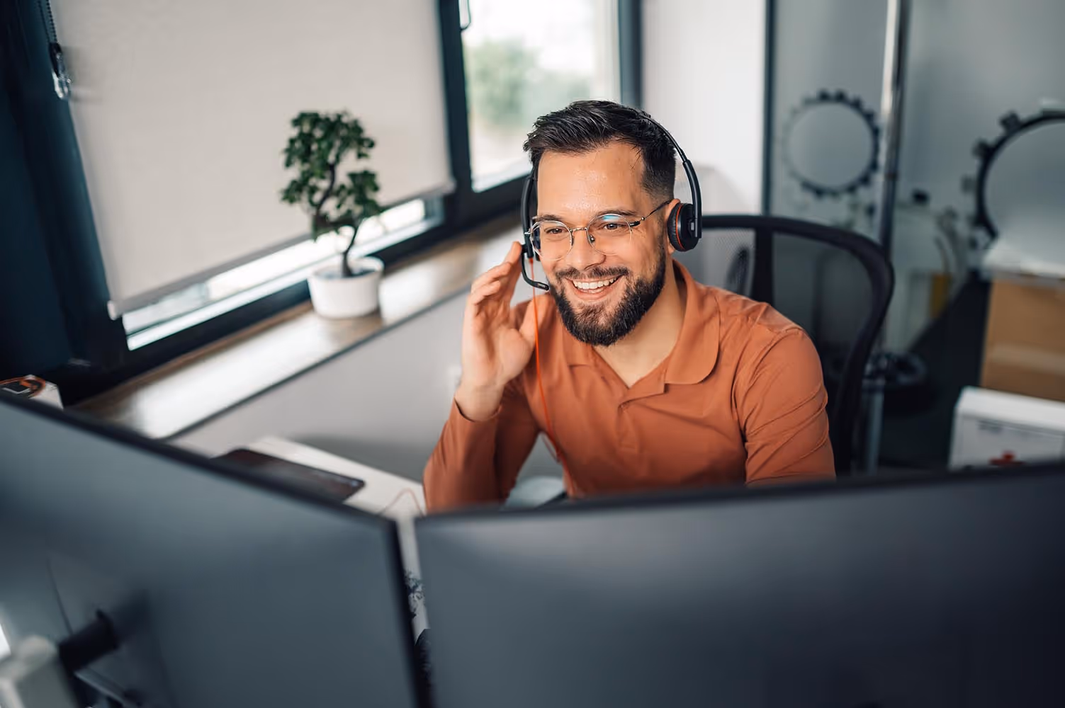 Smiling man wearing glasses and a headset, sitting at a desk with two computer monitors in a bright office.