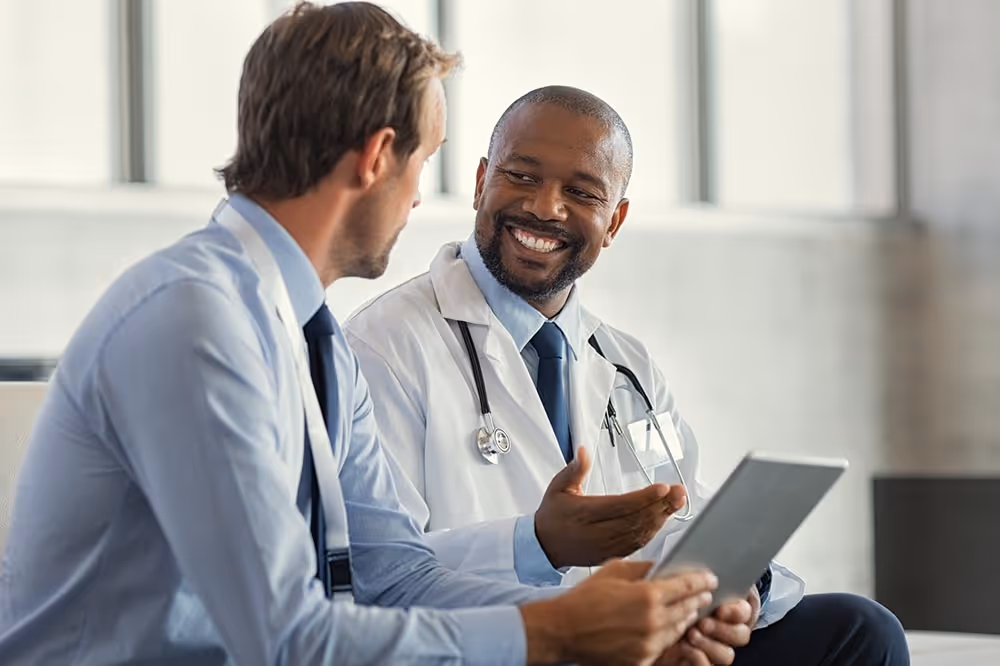 Two doctors in white coats smiling and discussing while looking at a tablet.