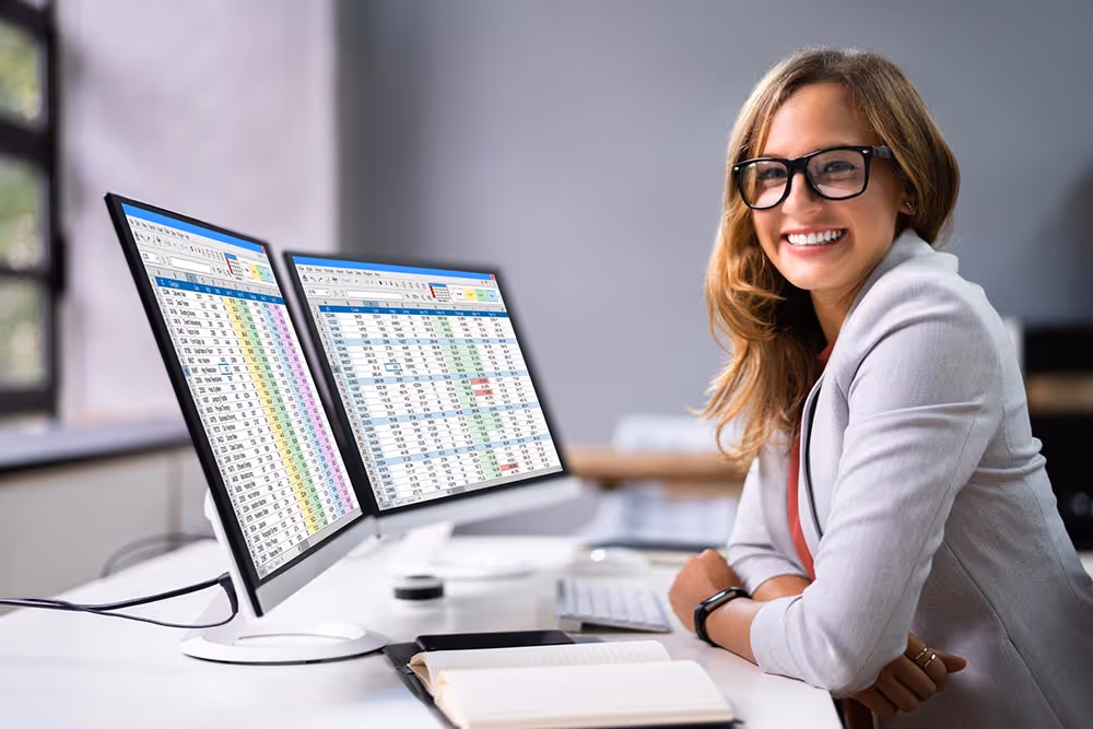 Smiling woman wearing glasses sitting at desk with two large monitors displaying spreadsheets.