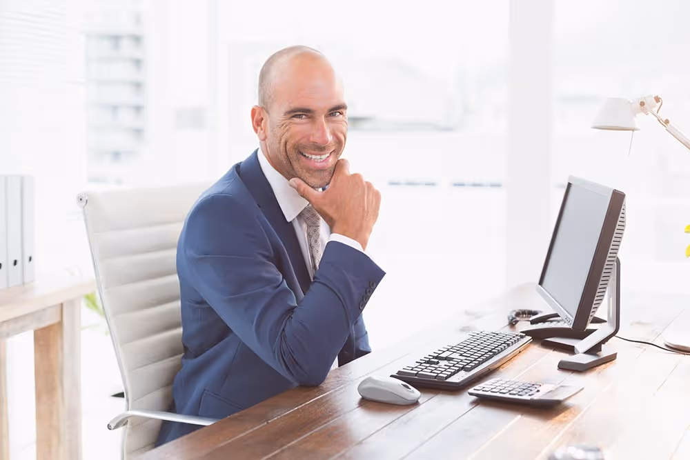 Smiling bald man in a blue suit sitting at a desk with a computer monitor, keyboard, mouse, and calculator in a bright office.