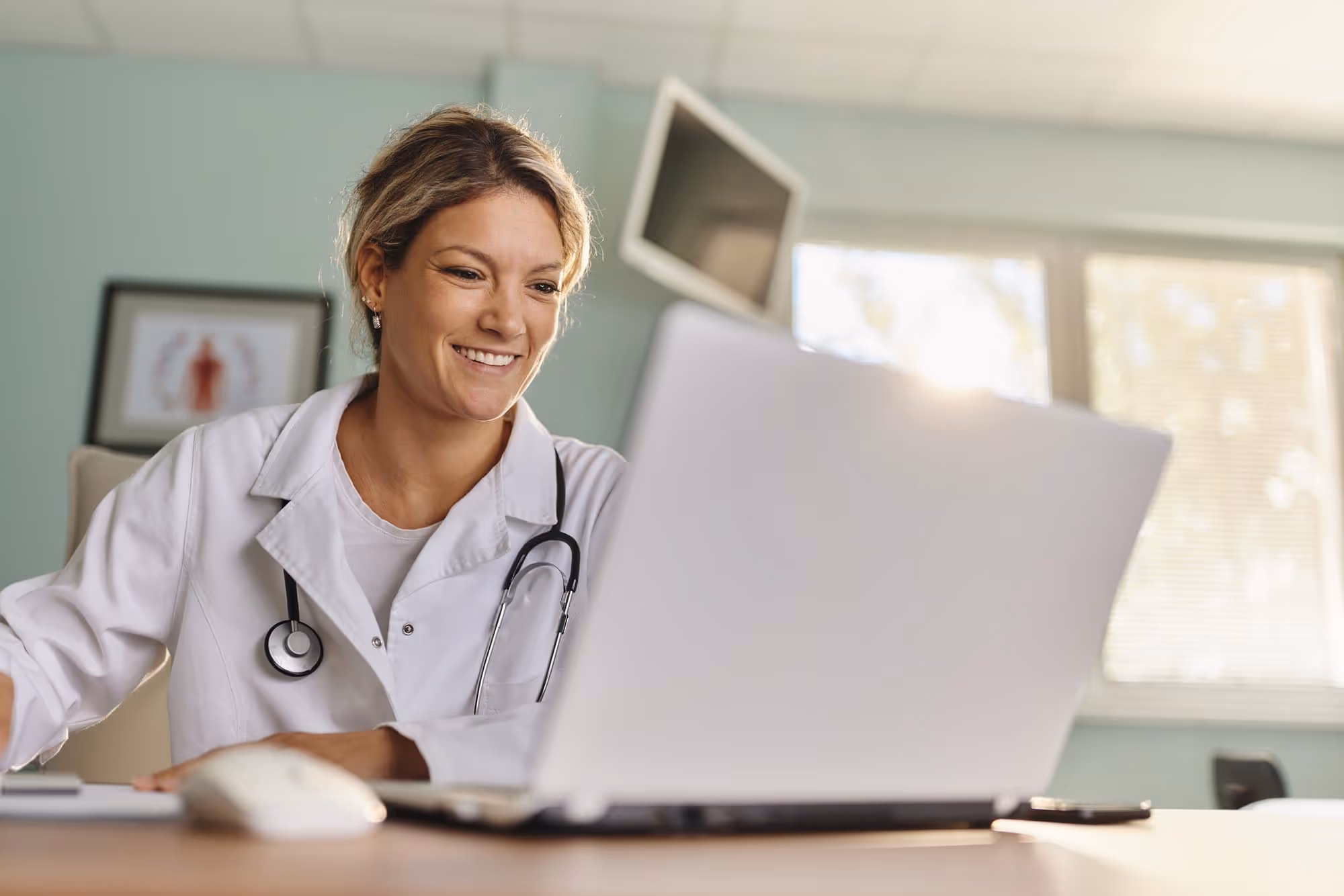 Smiling female doctor in white coat with stethoscope using a laptop in a bright office.