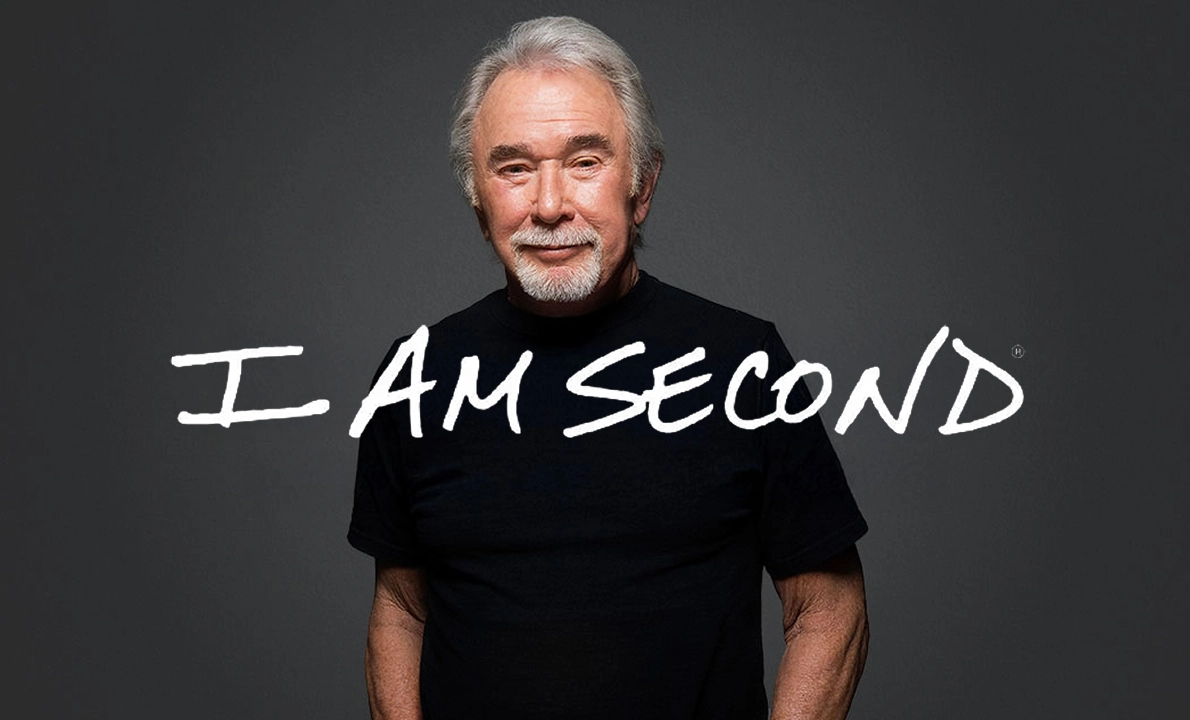 Older man with grey hair and beard wearing a black t-shirt standing in front of a dark background with white handwritten text saying I AM SECOND.