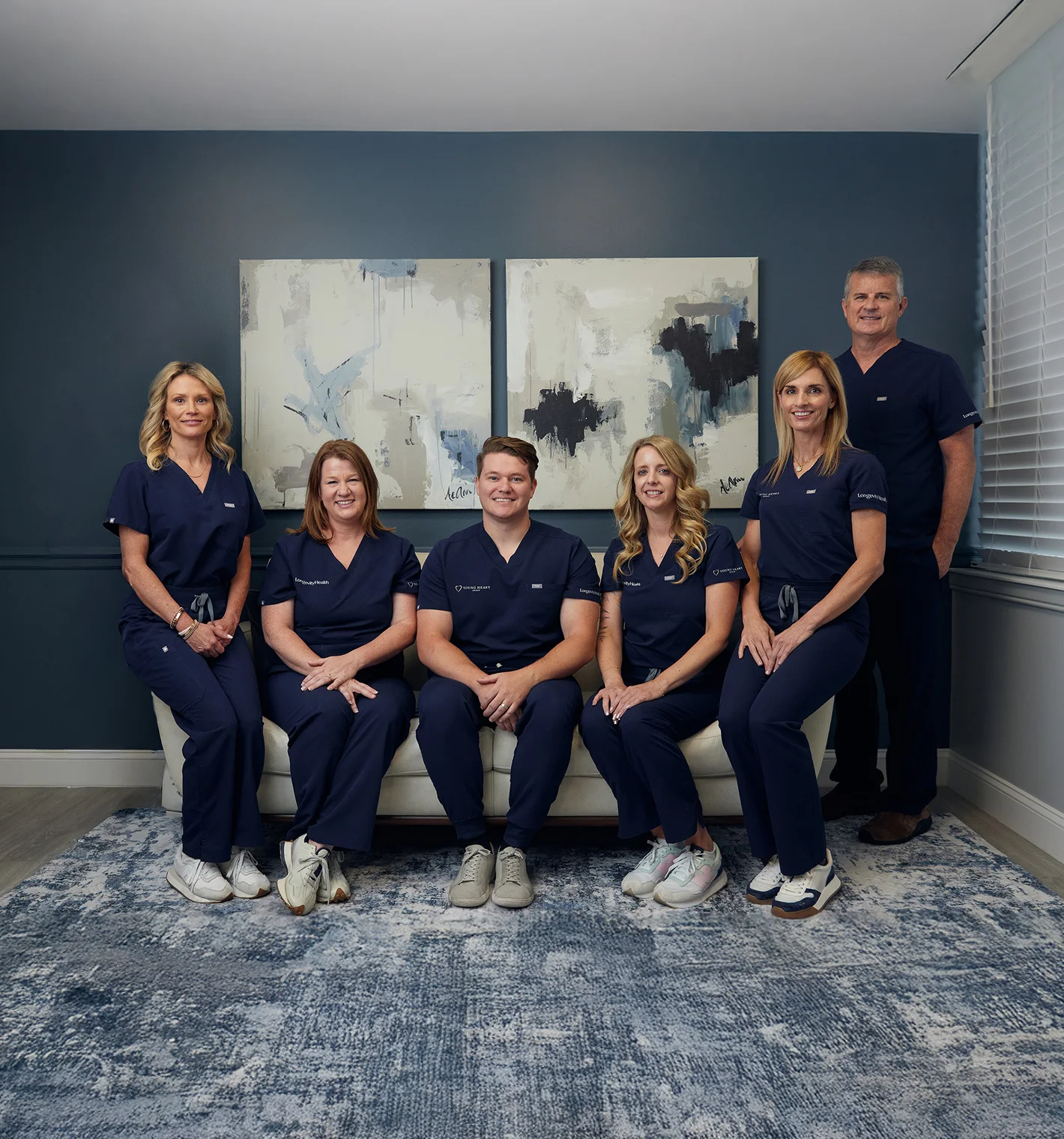 Six healthcare professionals wearing navy scrubs posing in a room with abstract paintings on the wall and a blue patterned rug.