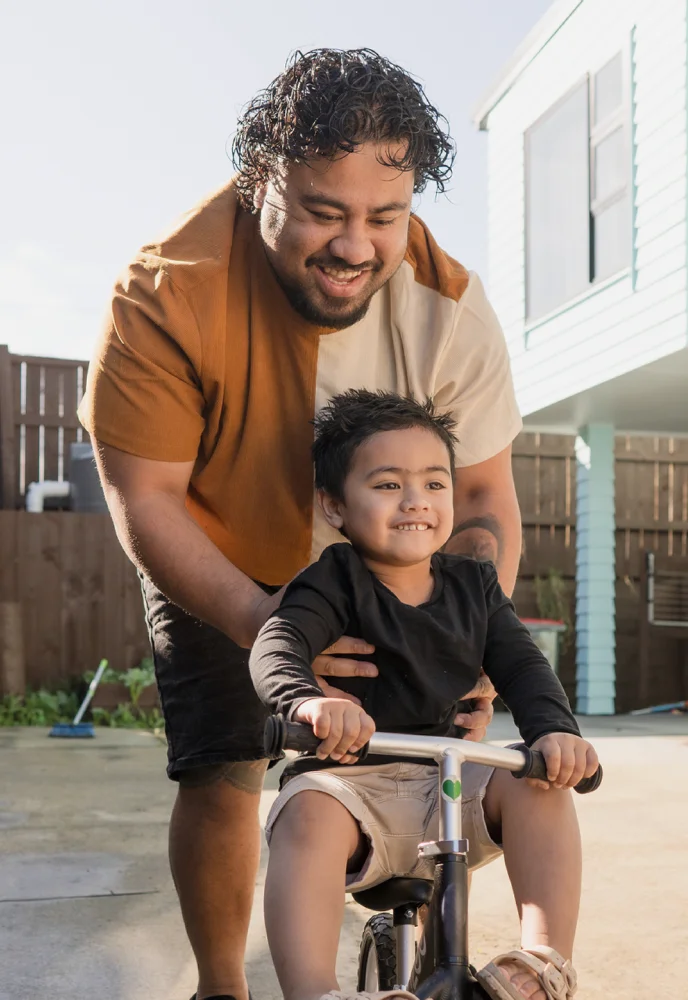 Smiling man helping young boy ride a balance bike outdoors near a house.