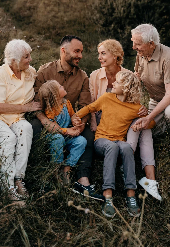Multi-generational family sitting outdoors in grass, smiling and holding hands, enjoying each other's company.