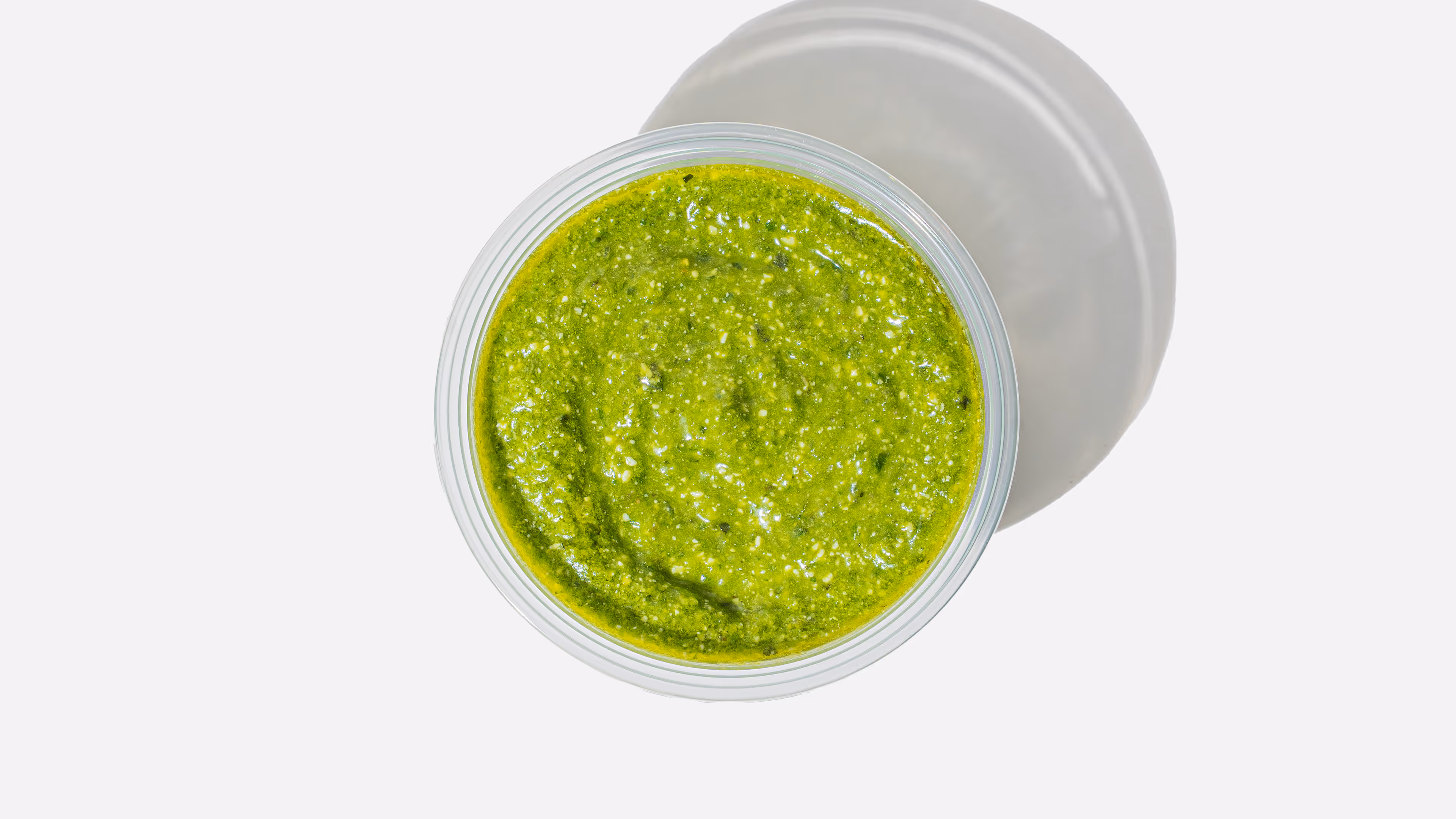 Top-down view of a clear plastic container filled with green pesto sauce casting a shadow on a white surface.