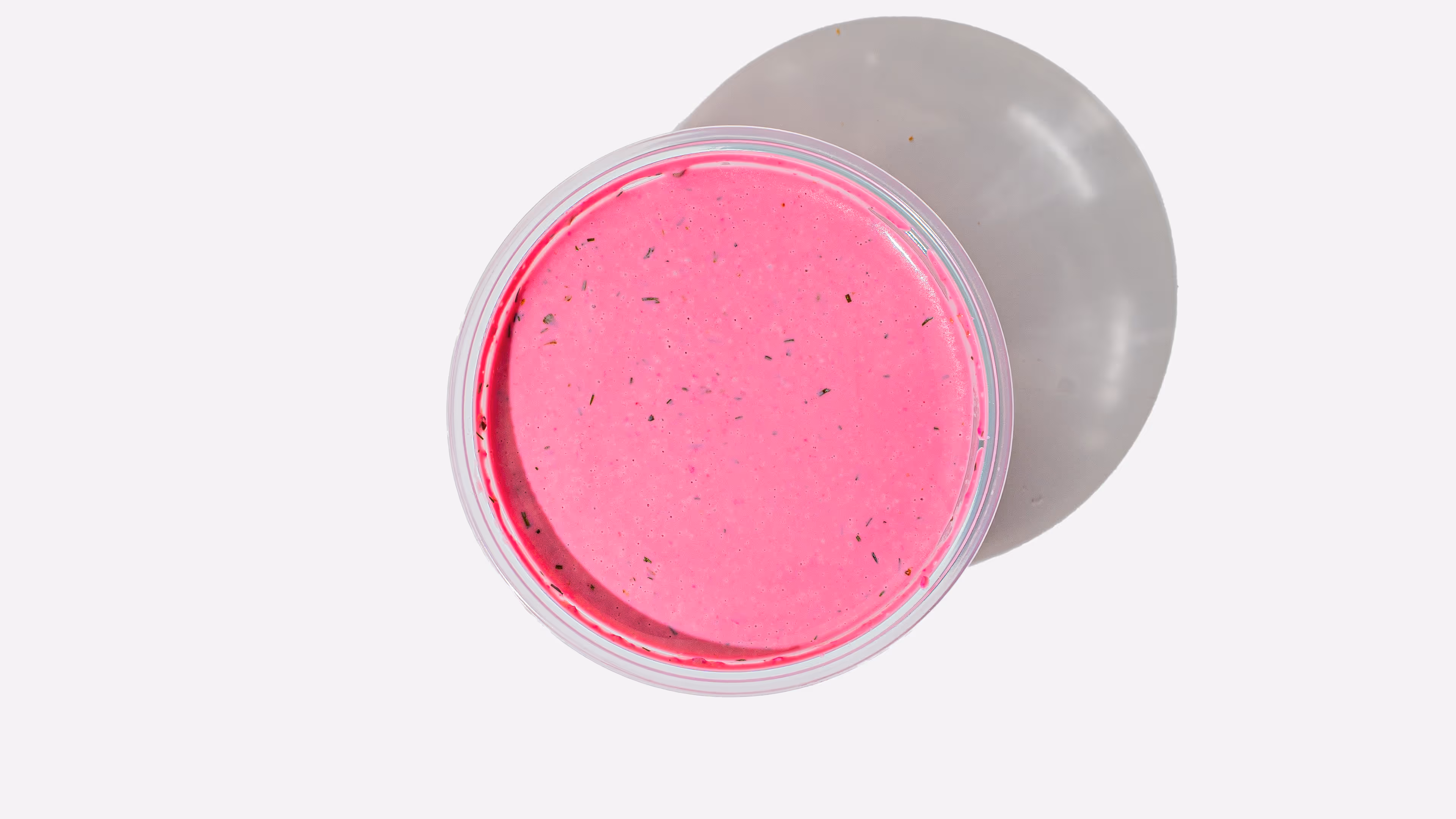 Top view of a clear round container filled with bright pink creamy sauce with herbs, casting a shadow on a white surface.