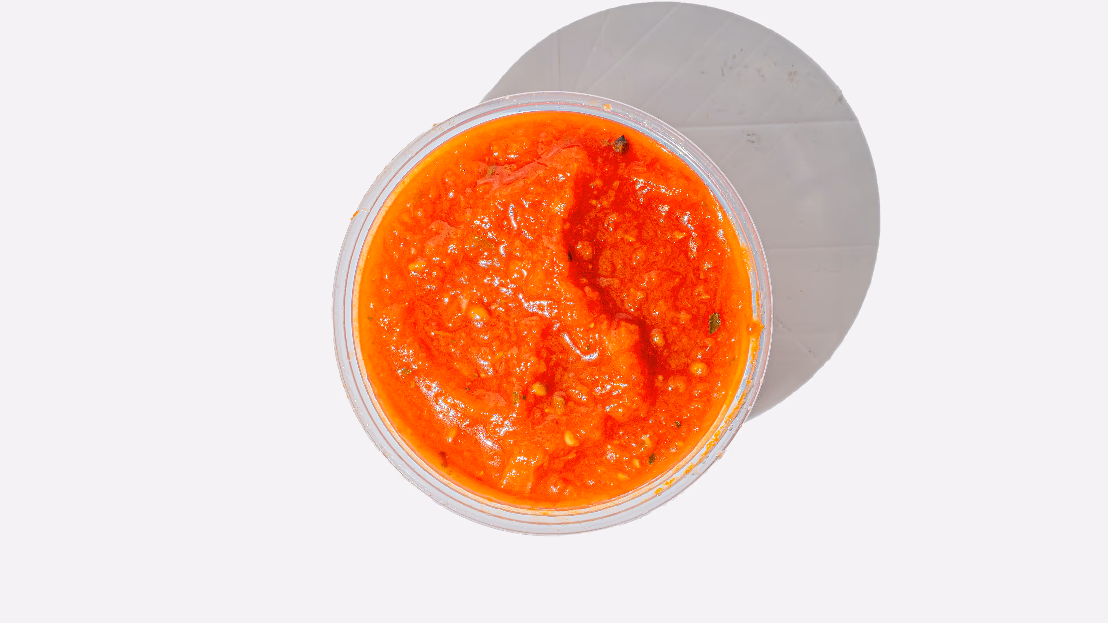 Top view of a round container filled with bright red chili sauce casting a shadow on a white surface.