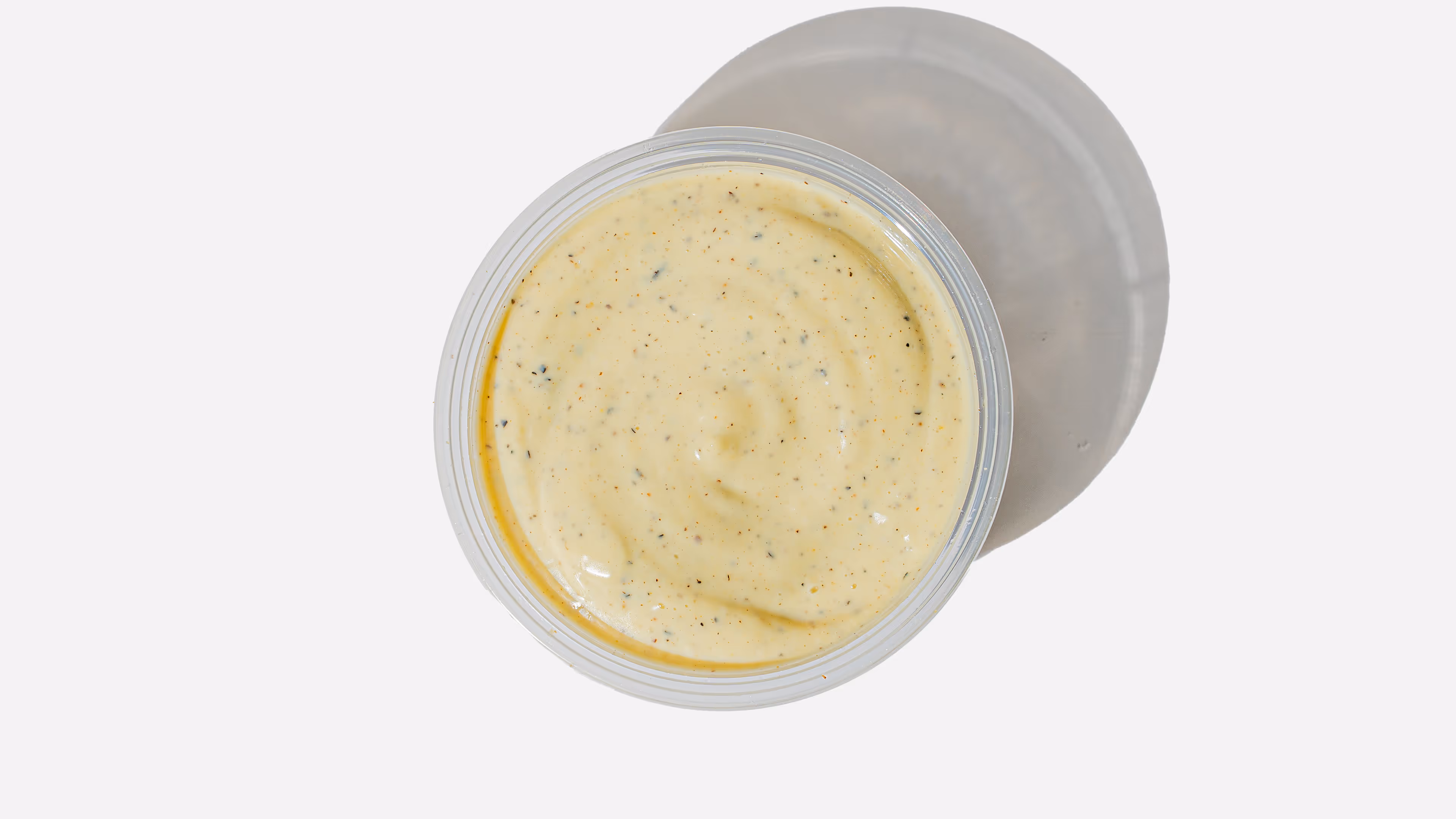 Top view of a plastic container filled with creamy yellow sauce speckled with herbs and spices casting a shadow.