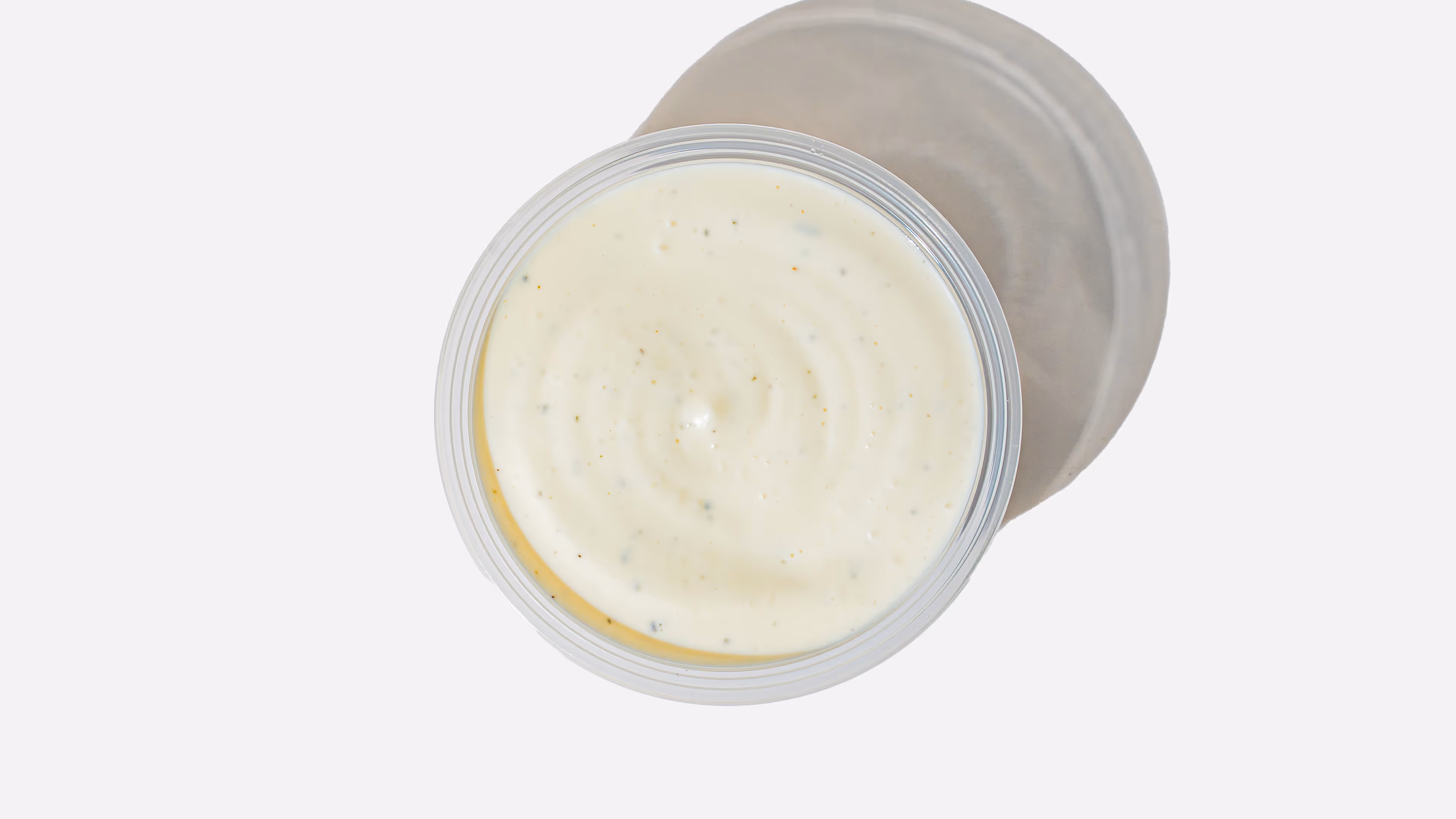 Top view of a clear plastic cup filled with creamy ranch dressing, casting a shadow on a white background.