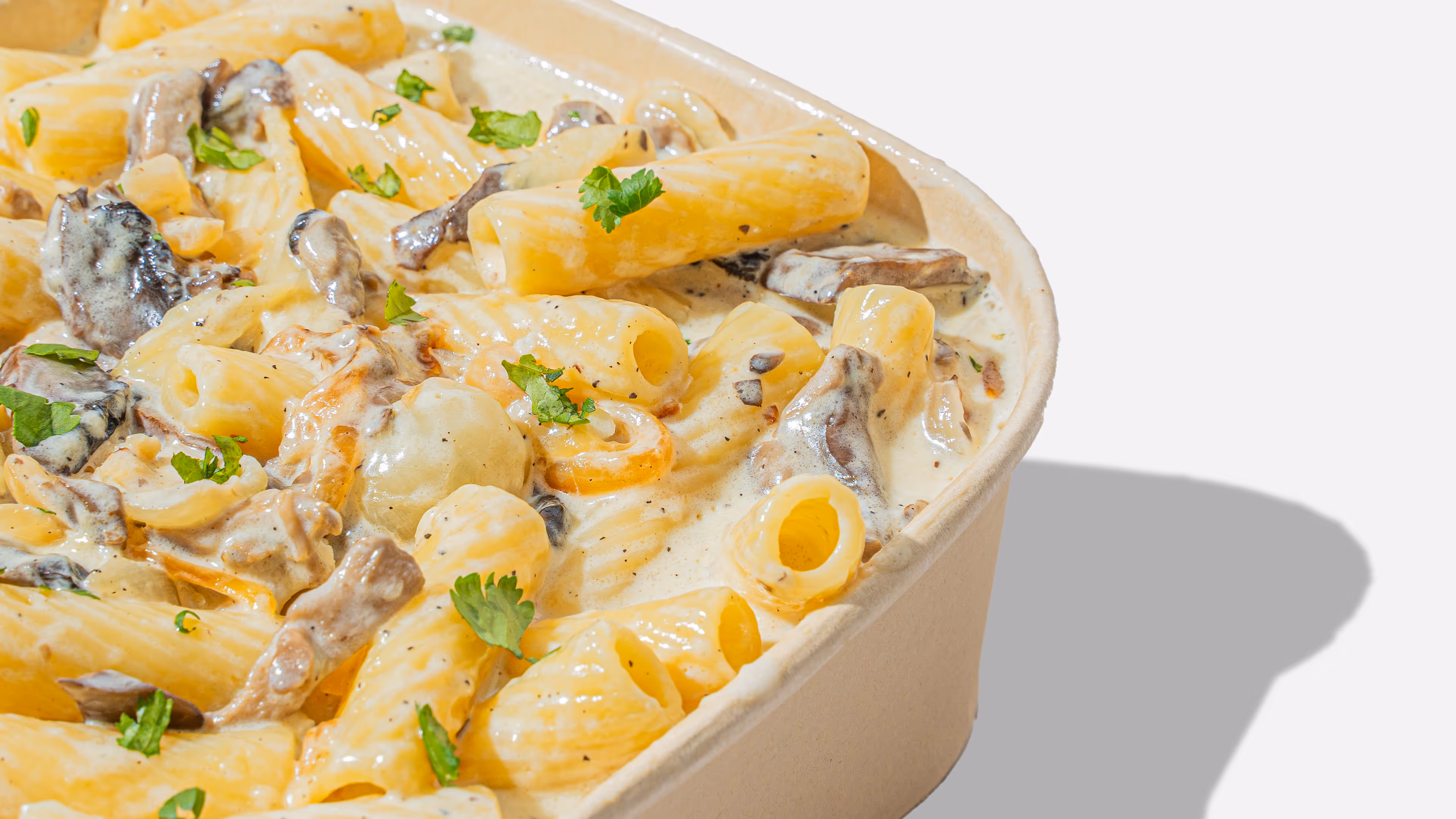 Close-up of creamy rigatoni pasta with mushrooms and parsley in a beige takeout container.