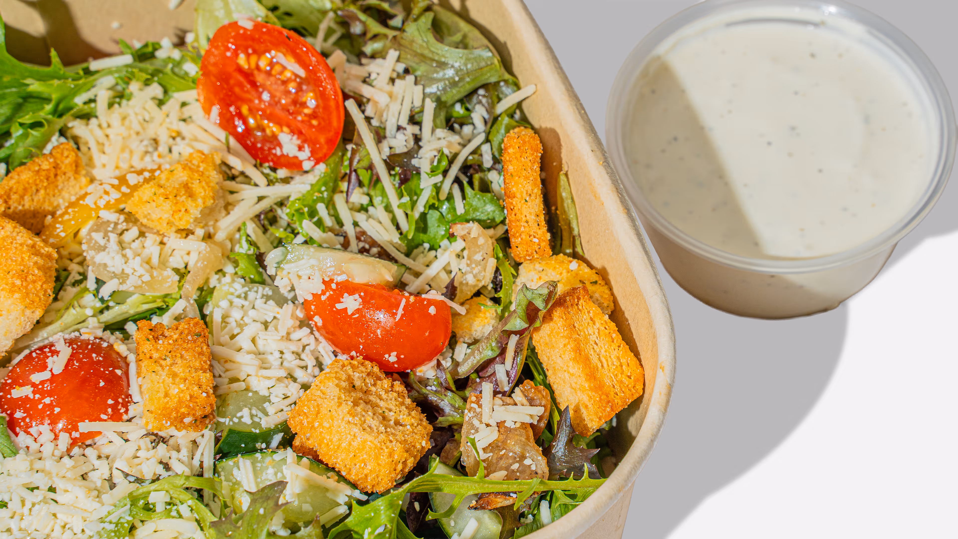 Fresh salad with cherry tomatoes, mixed greens, shredded cheese, croutons, and a small cup of creamy dressing.