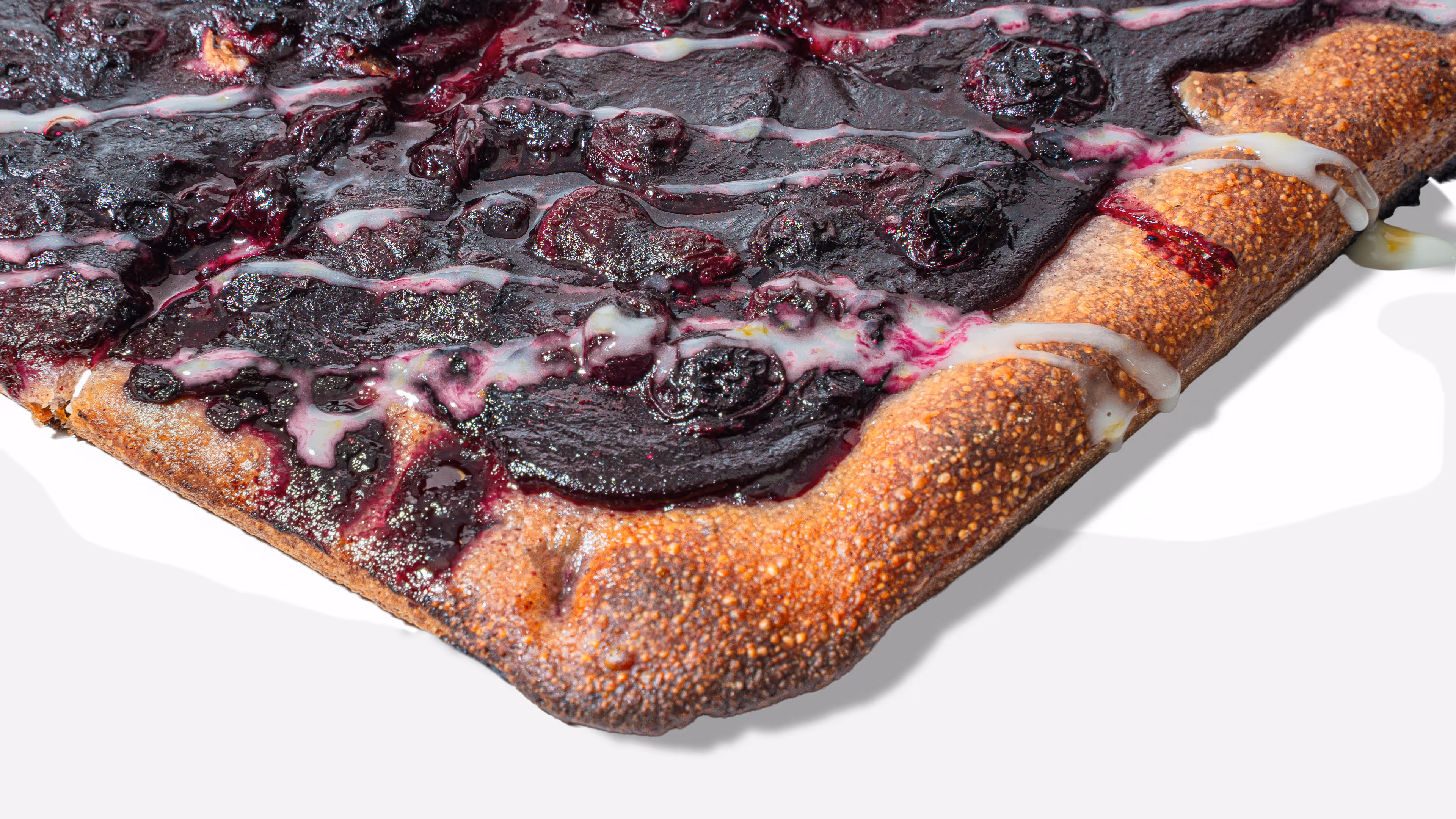 Close-up of a slice of blueberry dessert pizza with purple blueberry topping and white icing drizzle on a golden crust.
