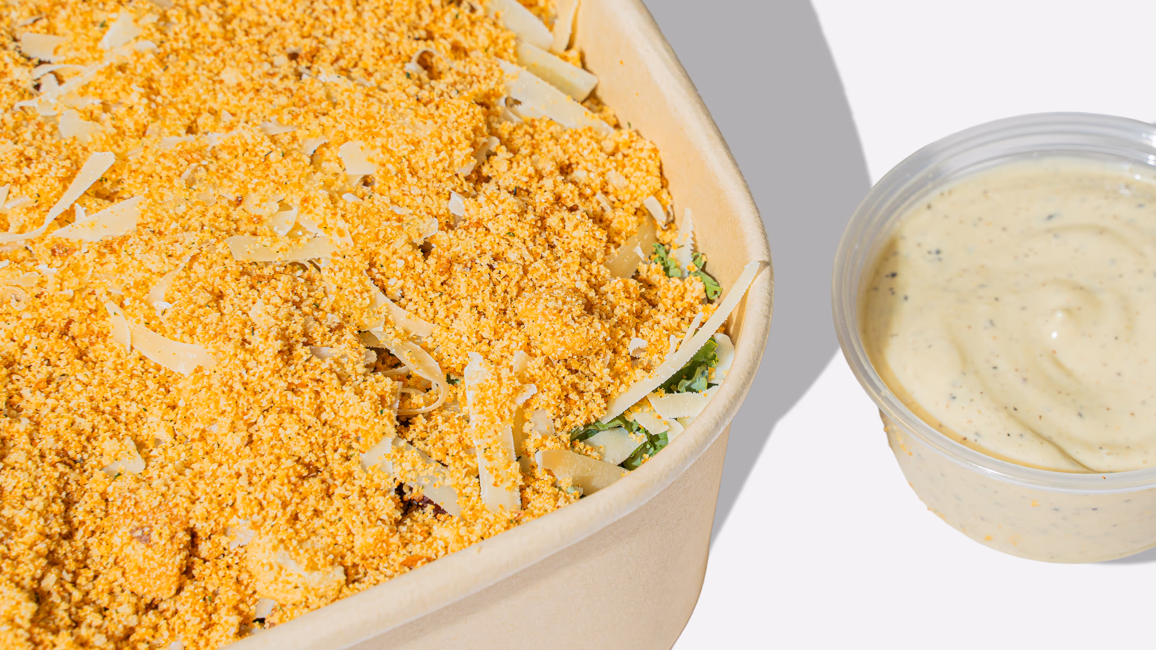 Close-up of a rectangular container filled with a leafy green salad topped with yellow grated cheese and a crispy crumb topping, accompanied by a small cup of creamy dressing.