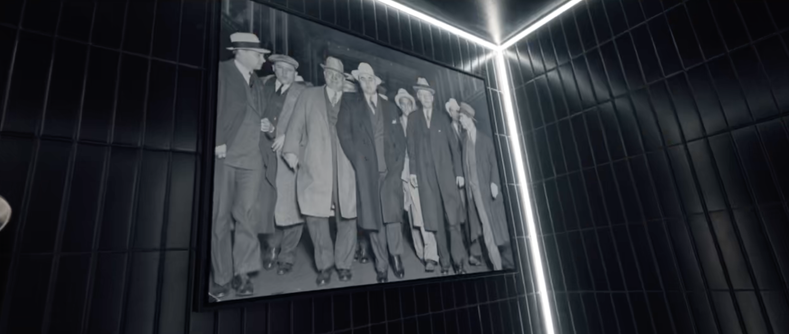 Black and white photo of a group of men in suits and hats walking together, displayed on a dark tiled wall with bright corner lighting.