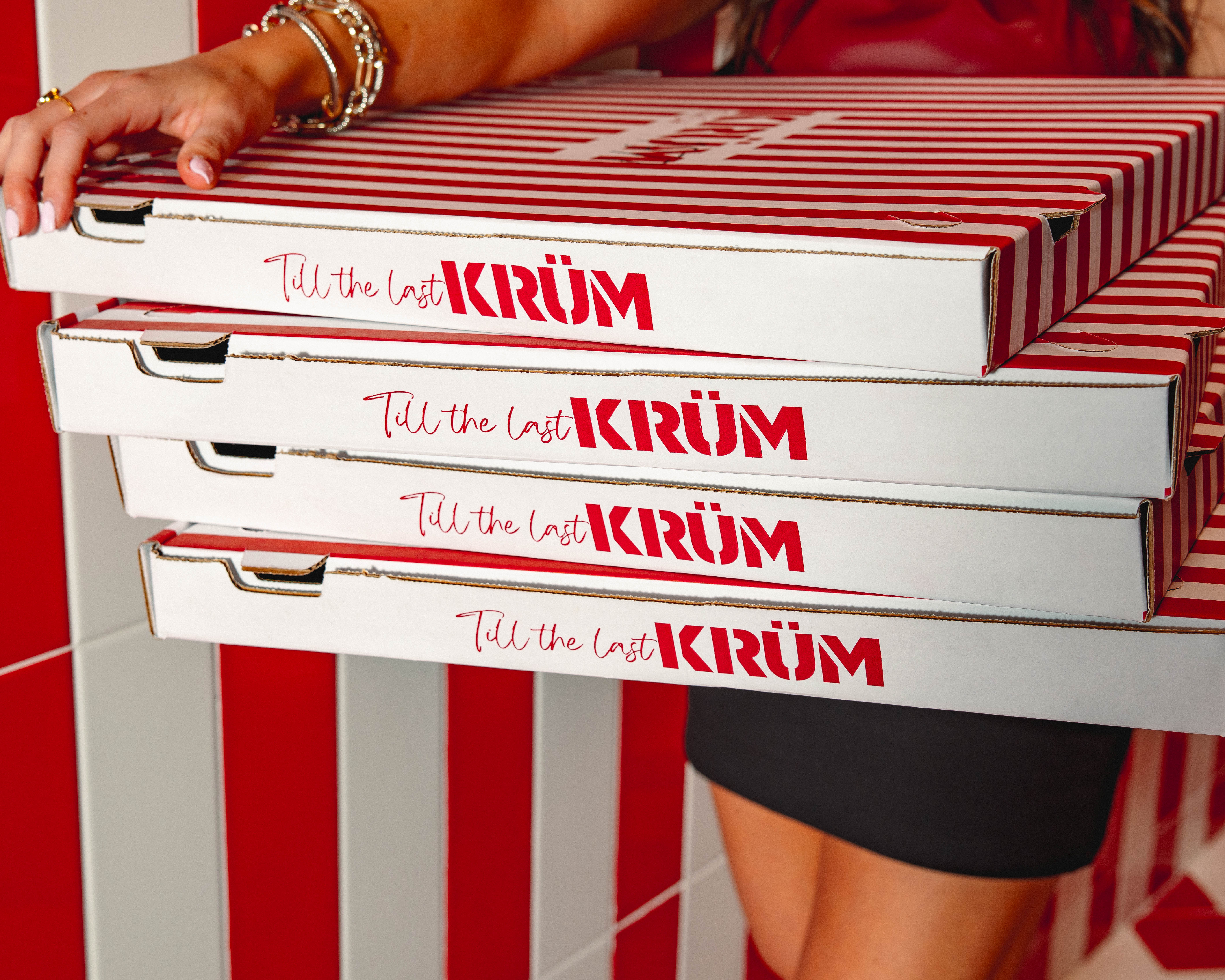 Person holding four stacked white and red striped pizza boxes with the text 'Till the last KRÜM' printed on the side.