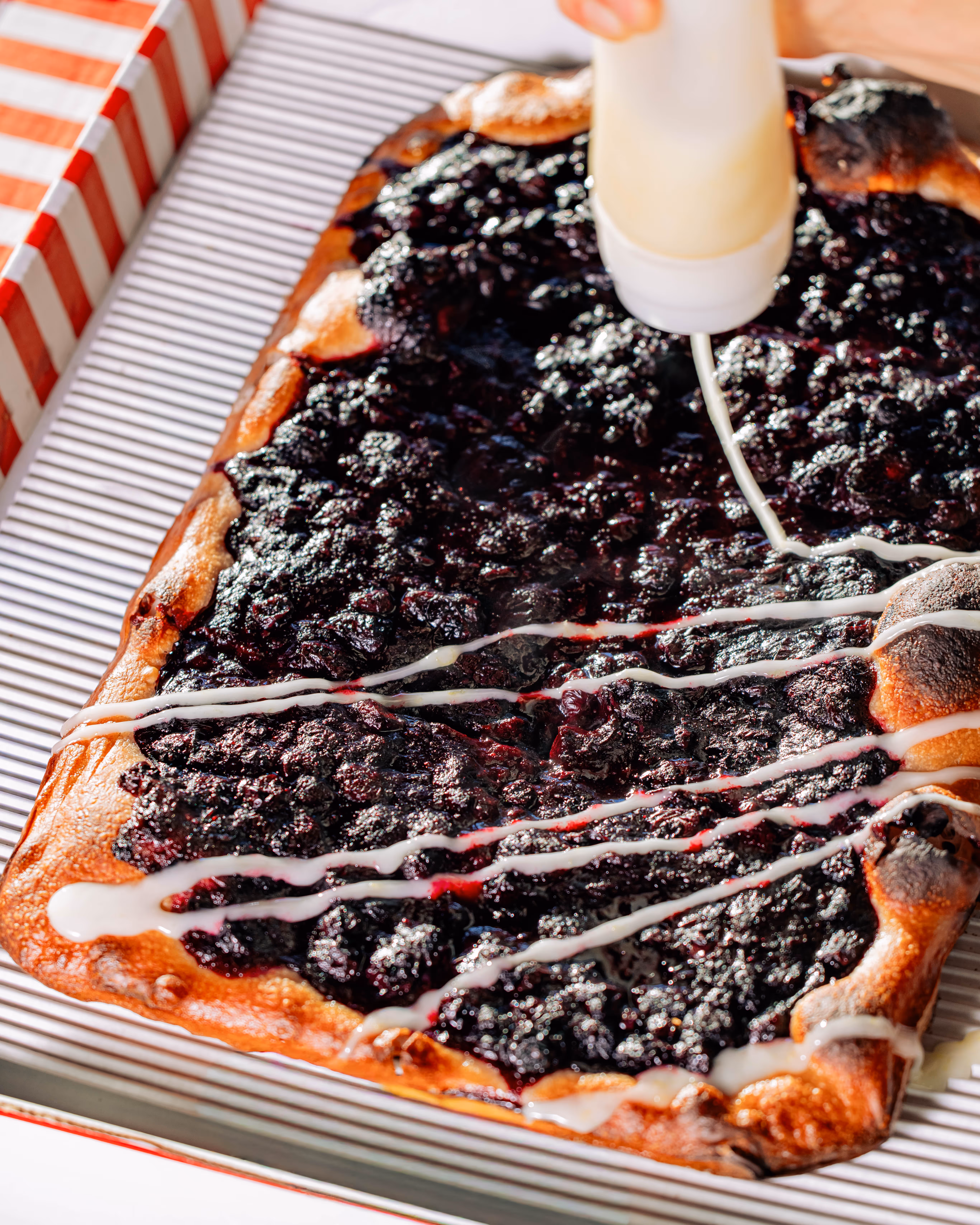 Close-up of a rectangular berry dessert with a golden crust being drizzled with white icing from a squeeze bottle.