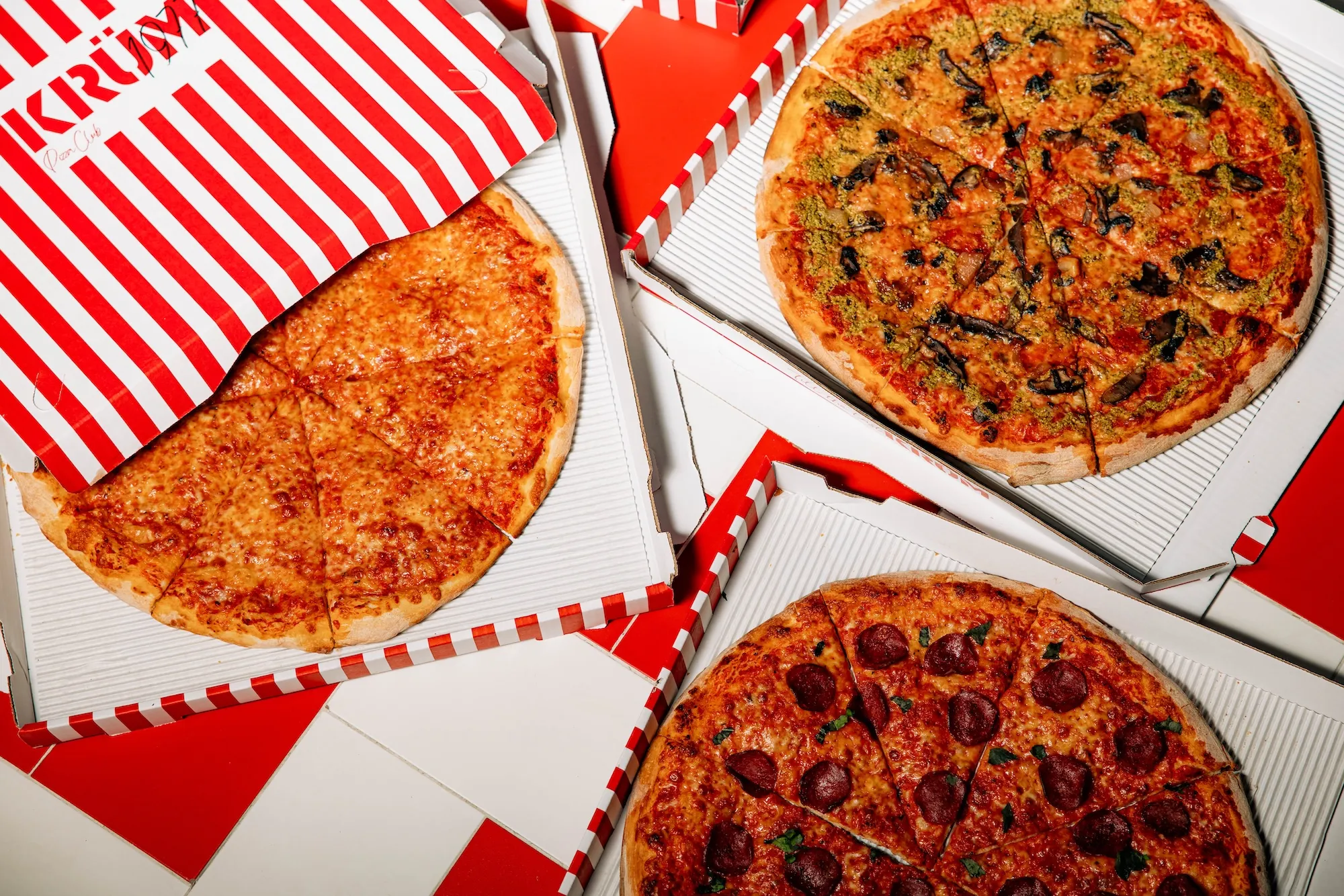 Three pizzas in open red and white striped boxes placed on red and white tiled floor, with cheese, mushroom pesto, and pepperoni toppings.