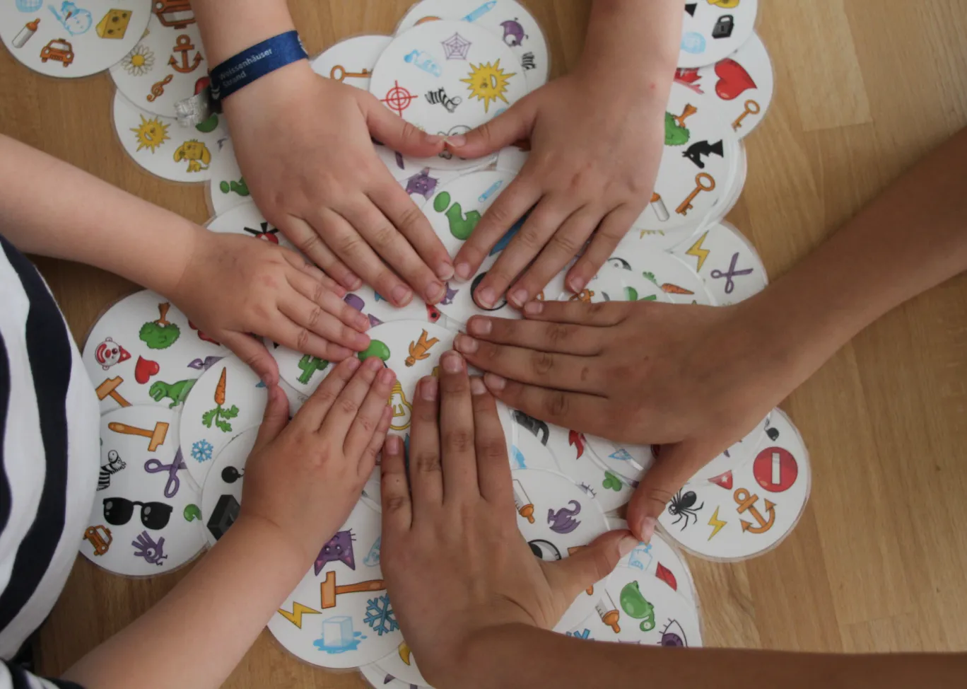 Fünf Kinderhände legen auf einem Holztisch mehrere runde Spielkarten mit bunten Symbolen.