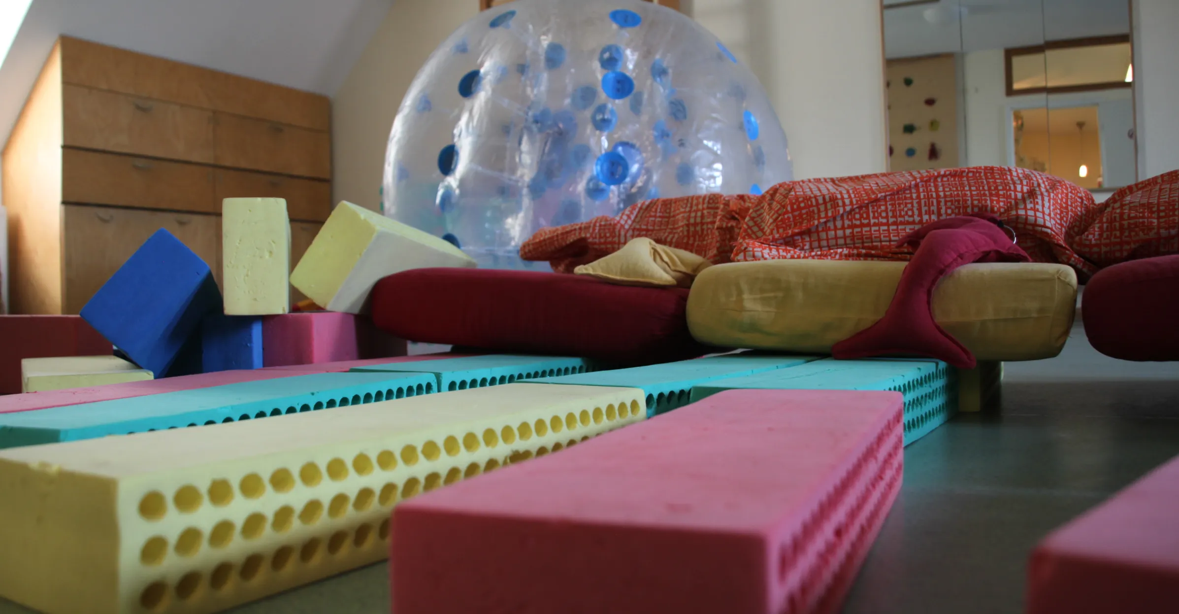 Bunte Schaumstoffmatratzen und Kissen liegen auf dem Boden und einem Sofa in einem Raum mit Holzmöbeln und einem großen durchsichtigen Ball mit blauen Punkten.