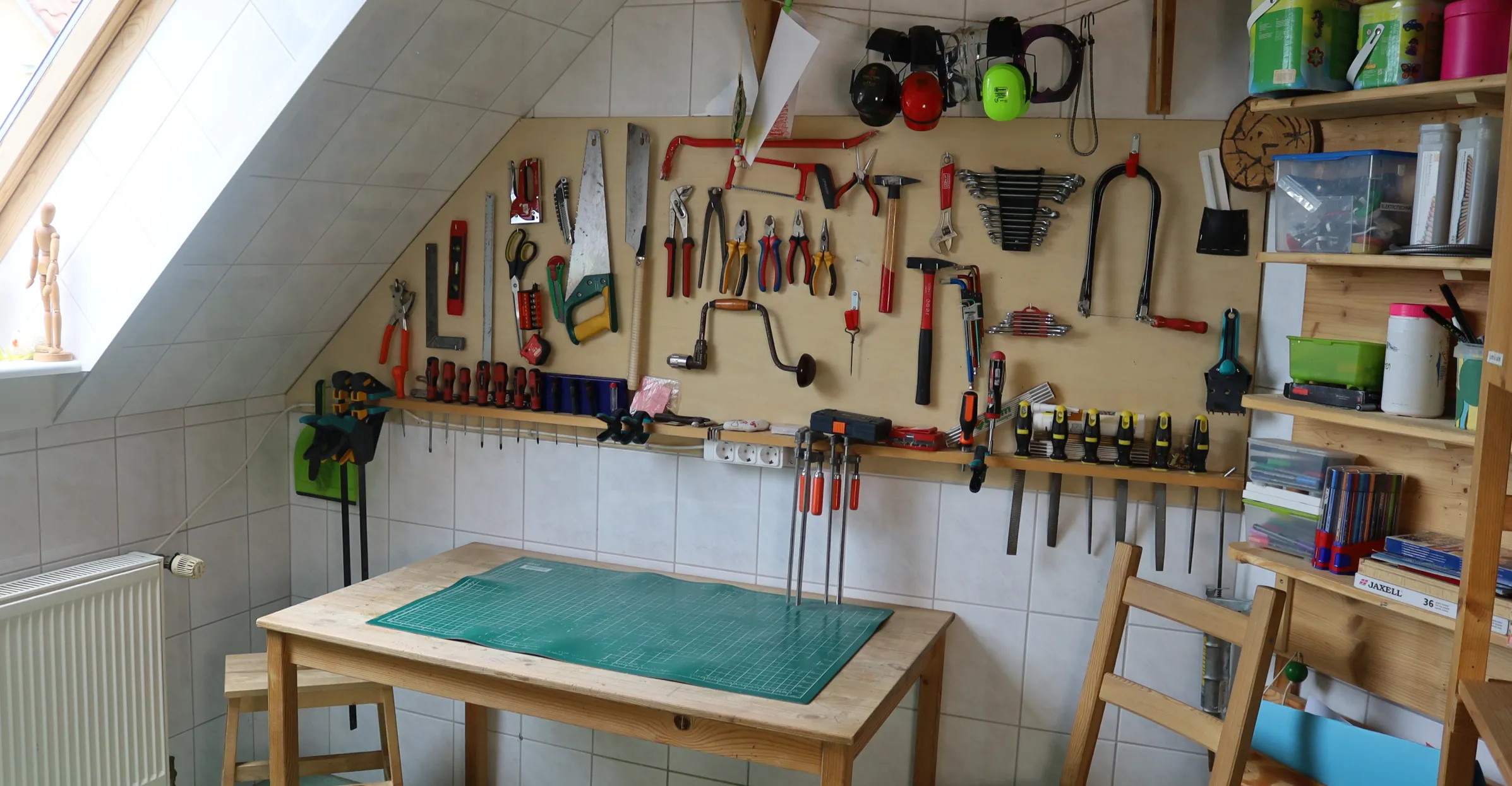 Werkstatt mit Holzarbeitstisch, Werkzeugwand mit Sägen, Schraubenzieher, Zangen und Hämmern, Regal mit Behältern und Stühlen.