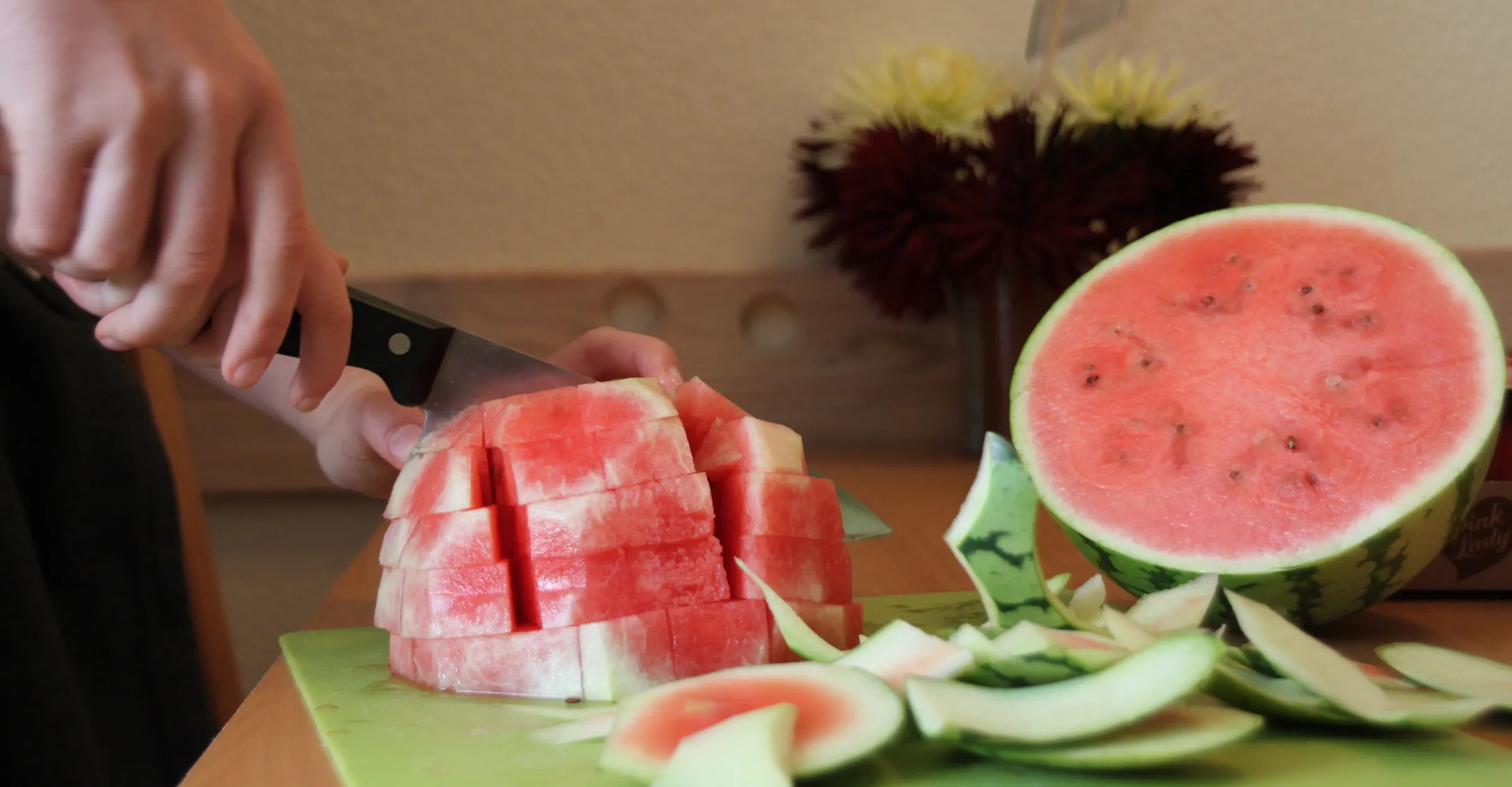 Person schneidet eine gewürfelte Wassermelone auf einem Schneidebrett, daneben liegen Melonenschalen und eine halbe Melone.
