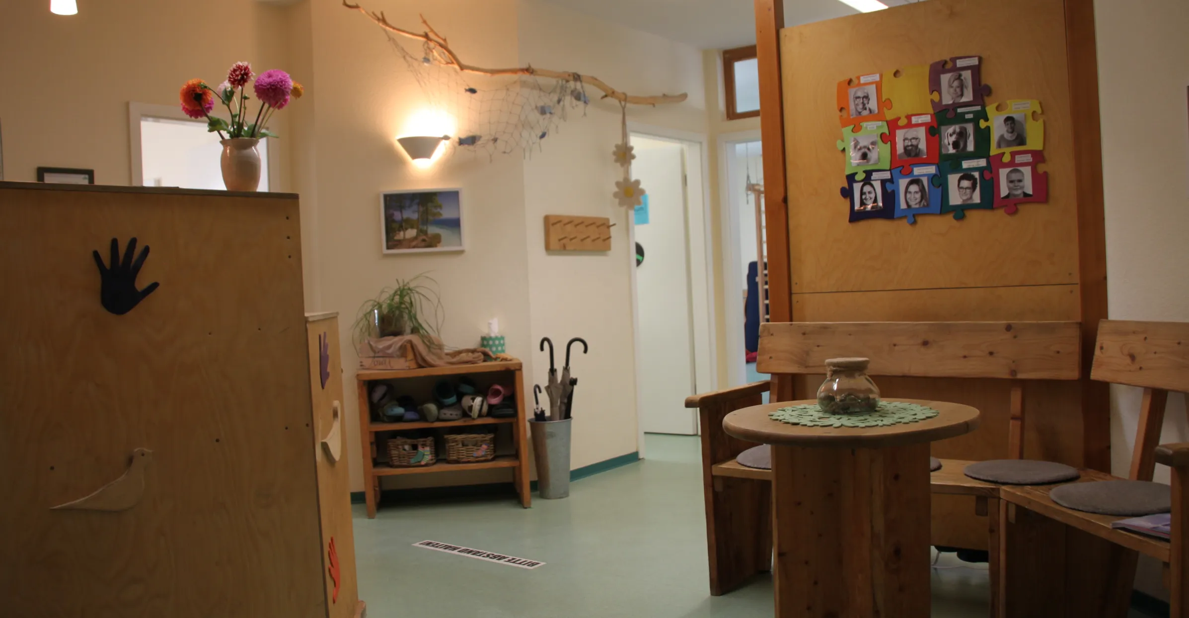 Wartebereich mit Holzmöbeln, einem runden Tisch, einer Vase mit Blumen und Fotos an der Wand.