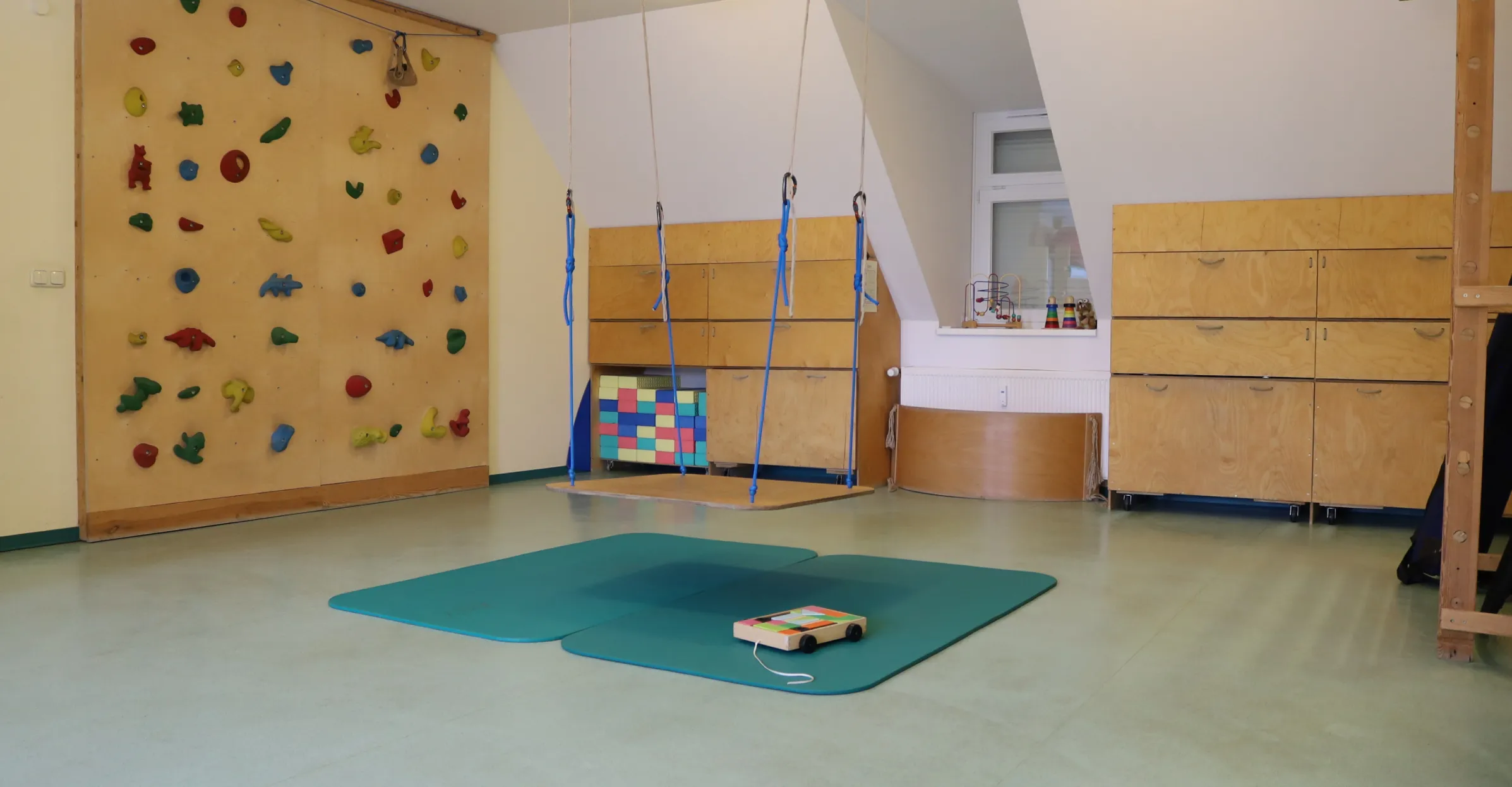 Raum mit Kletterwand, hängender Schaukel, blauen Matten auf dem Boden und Holzschränken mit Spielzeug im Fenster.