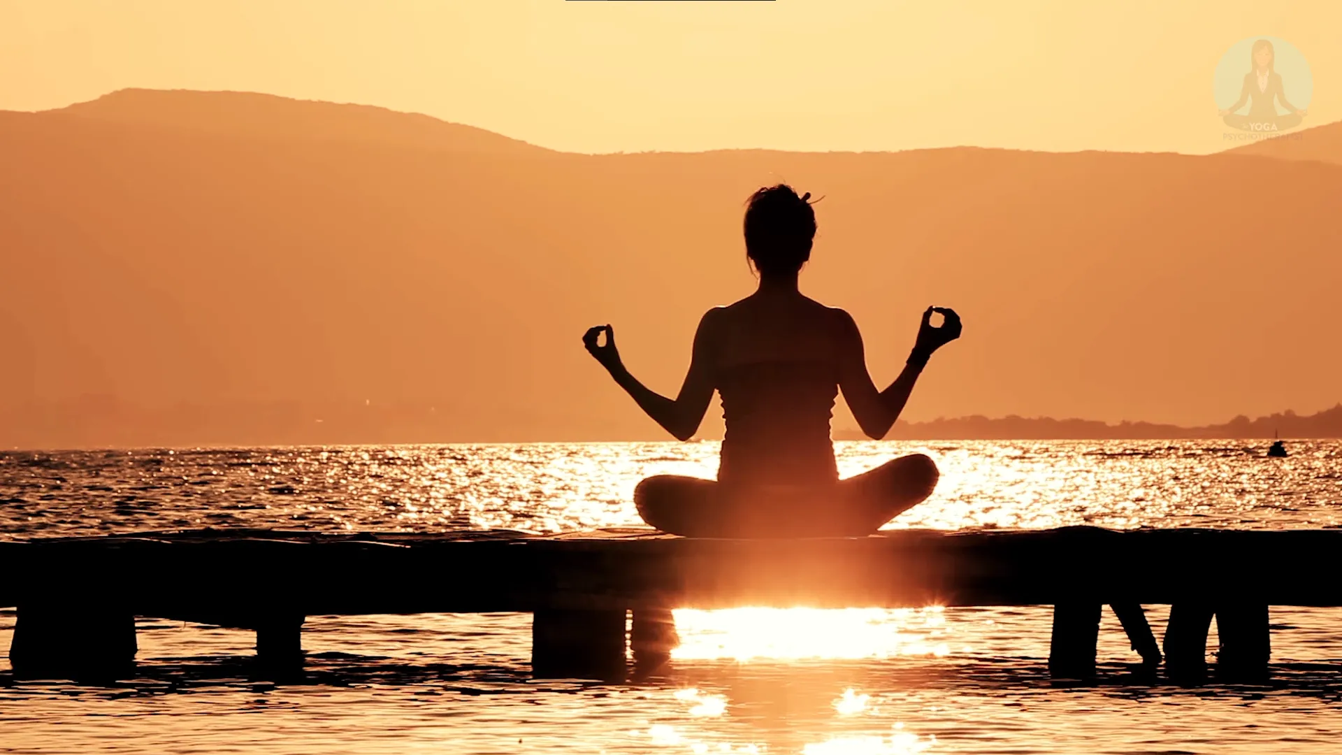 Silhouette of a person meditating in lotus pose on a dock with a calm lake and mountains during sunset.